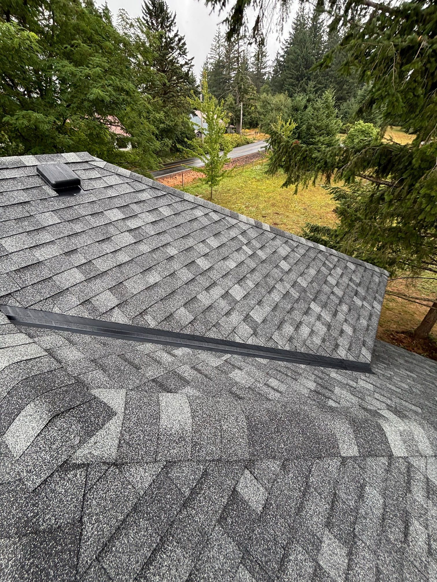 High-angle view of a gray asphalt shingled roof with a vent, transition flashing, and a background of green trees.