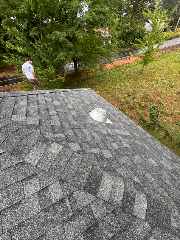 A person stands on a grey shingled roof next to a metal roof vent, overlooking a grassy yard with trees.