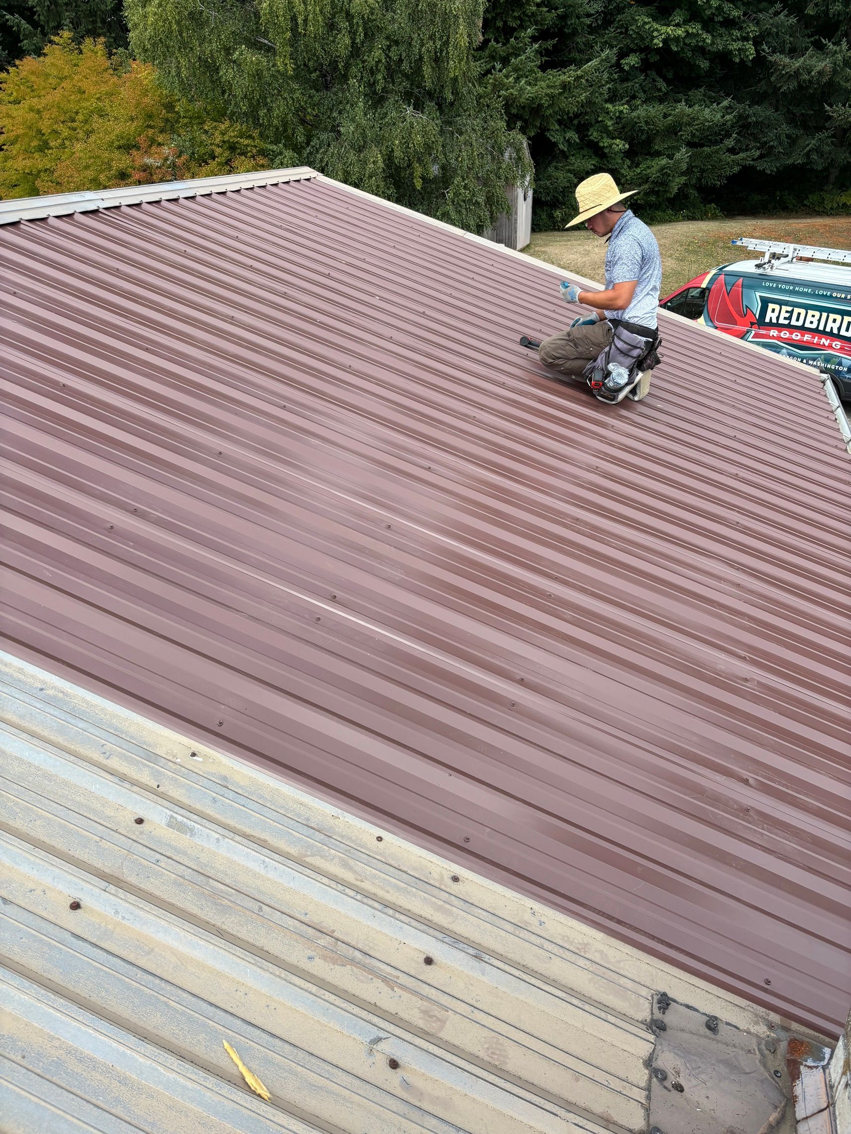 A worker wearing a straw hat installs reddish-brown corrugated metal roofing panels on an outdoor structure.