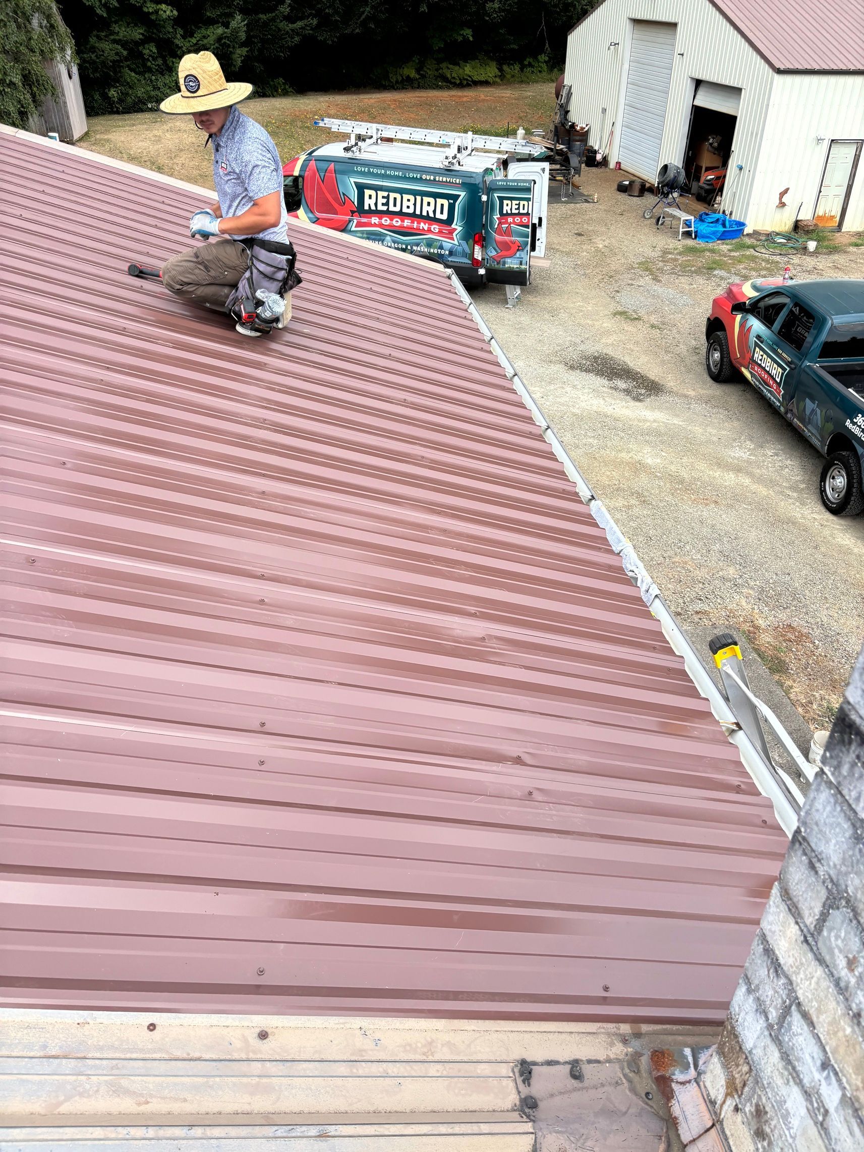 A worker in a hat kneels on a sloped, red metal roof, with a trailer and a pickup truck visible in the yard below.