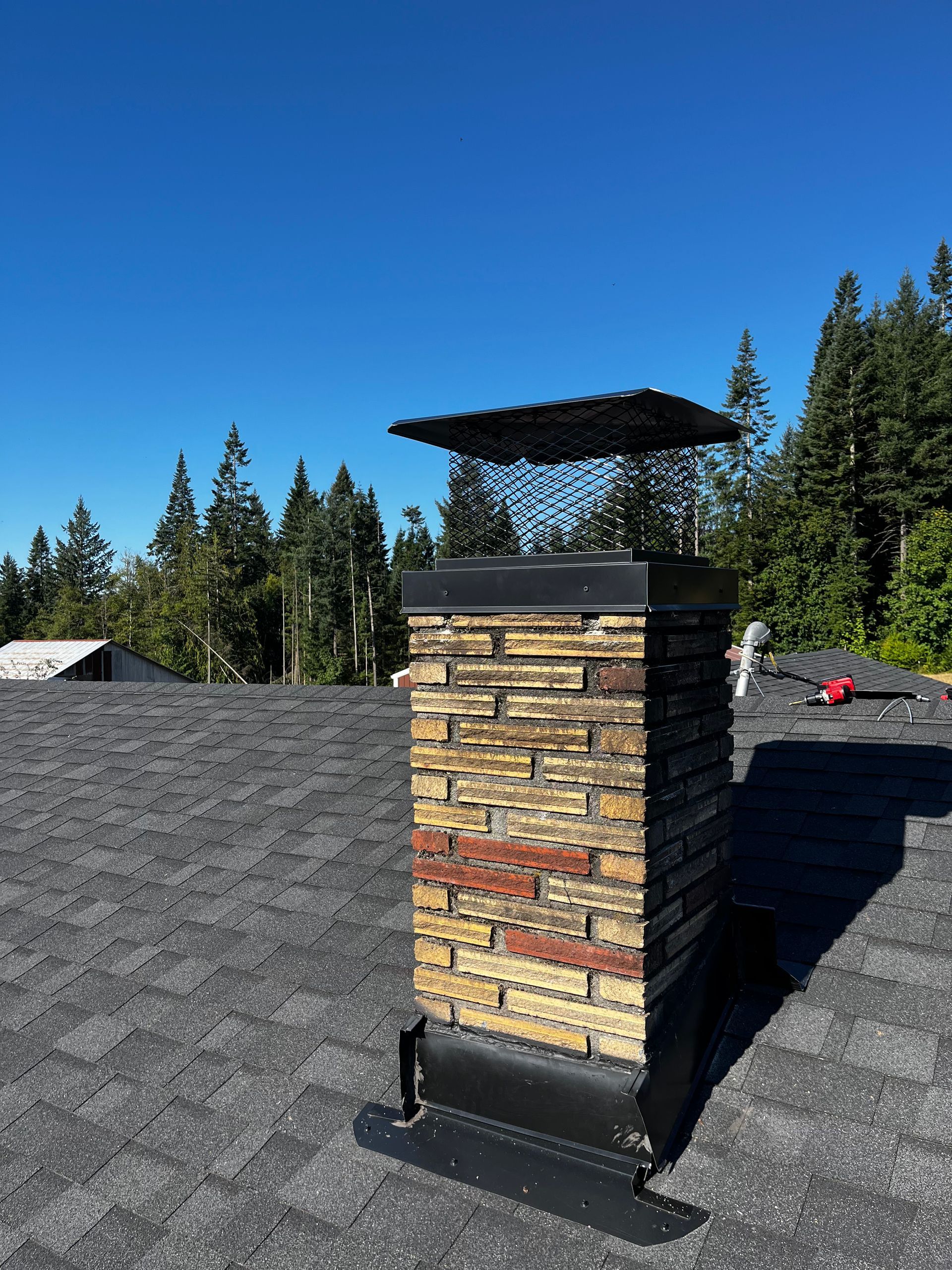 A brick chimney with a metal cap and flashing sits on a dark shingled roof under a clear blue sky and pine trees.