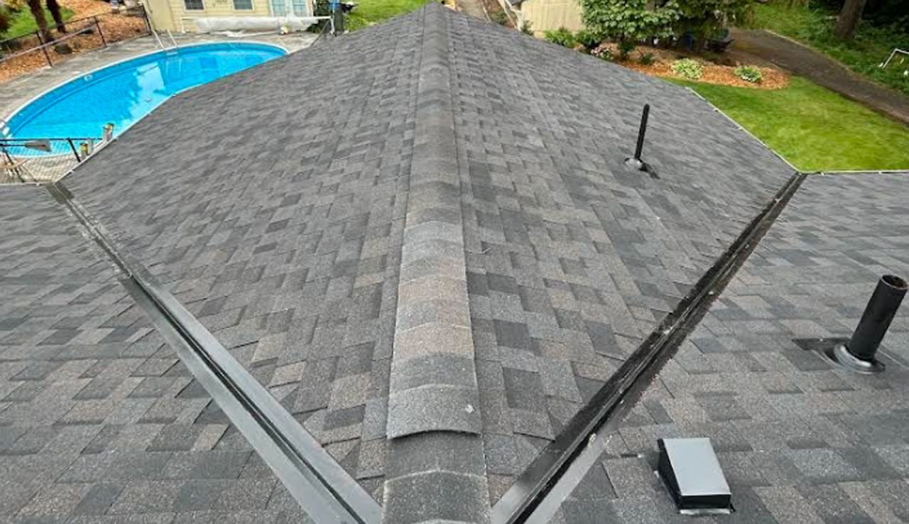 A high-angle view of a grey shingled roof with dark metal flashing in the valleys and a swimming pool in the background.