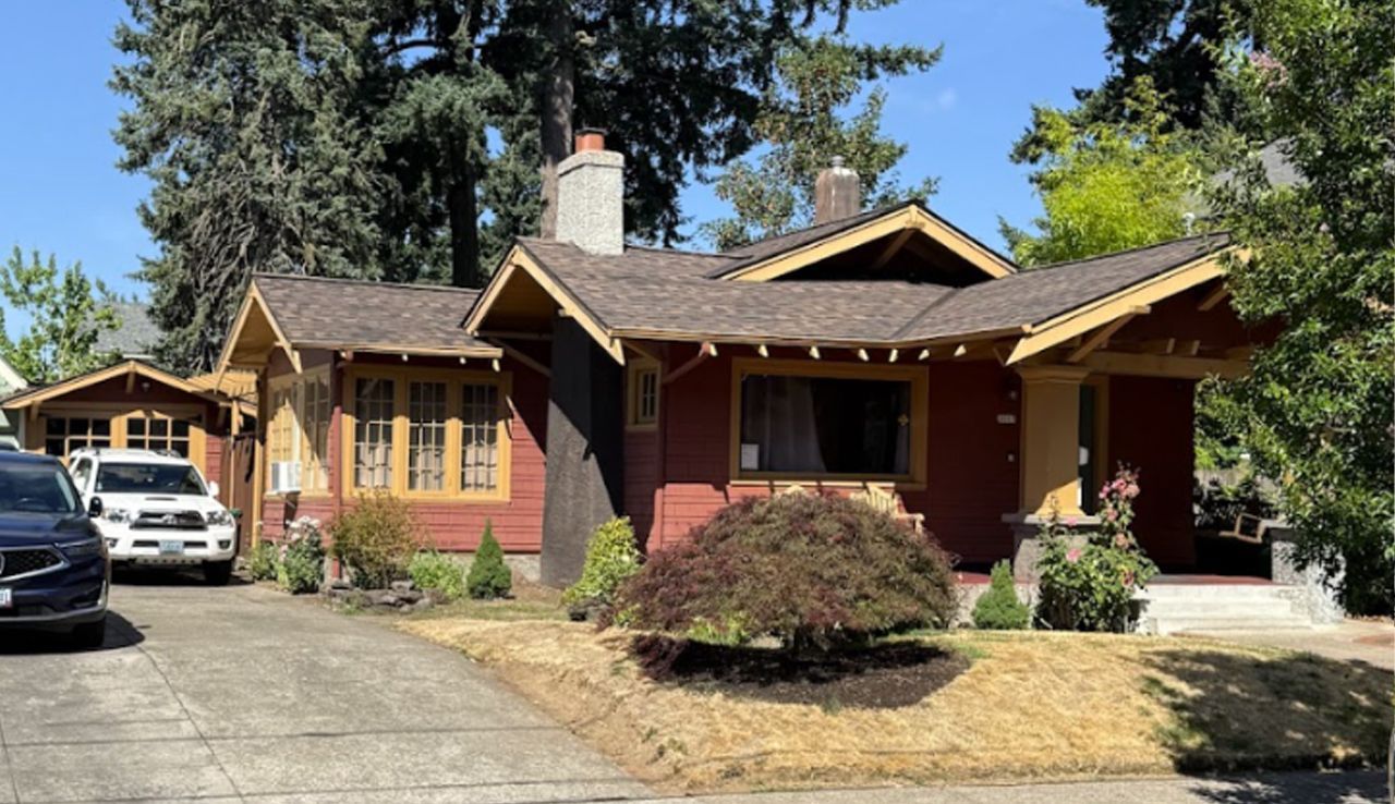 A red craftsman-style bungalow with a brown roof and a front porch, featuring a driveway with two parked vehicles.