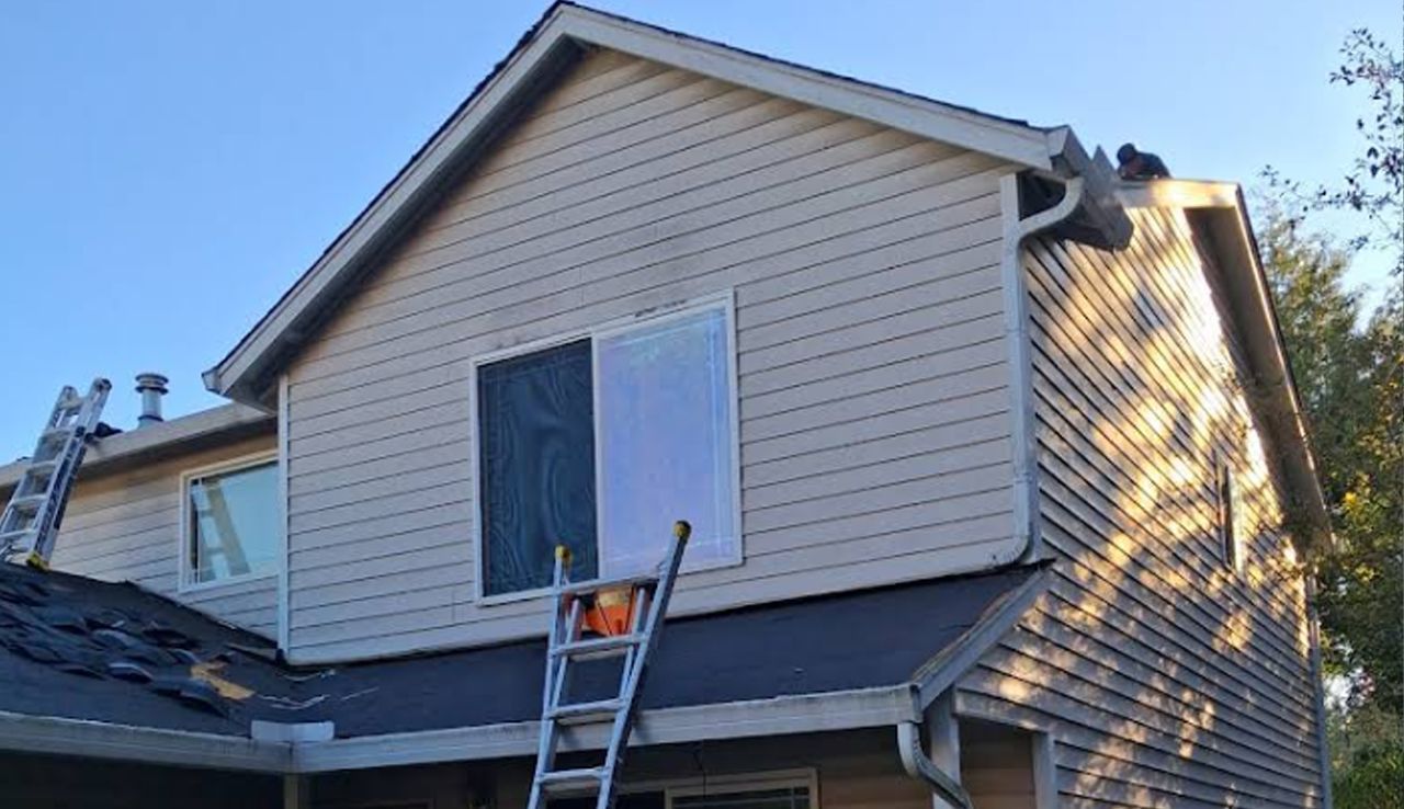A two-story house exterior under construction, showing roof repairs, a ladder, and a window with one side covered.