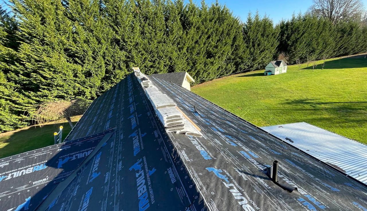 A rooftop under construction covered in black synthetic underlayment, with a bright green lawn and tall pine trees nearby.