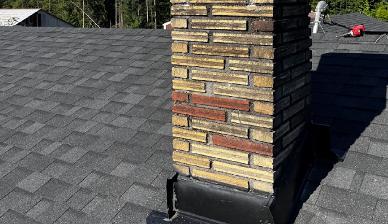 A brick chimney with mixed red and yellow bricks stands on a gray shingled roof with black flashing at the base.