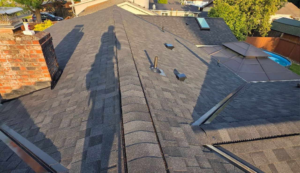 A high-angle view of a residential asphalt shingle roof with a brick chimney, metal vents, and a skylight.