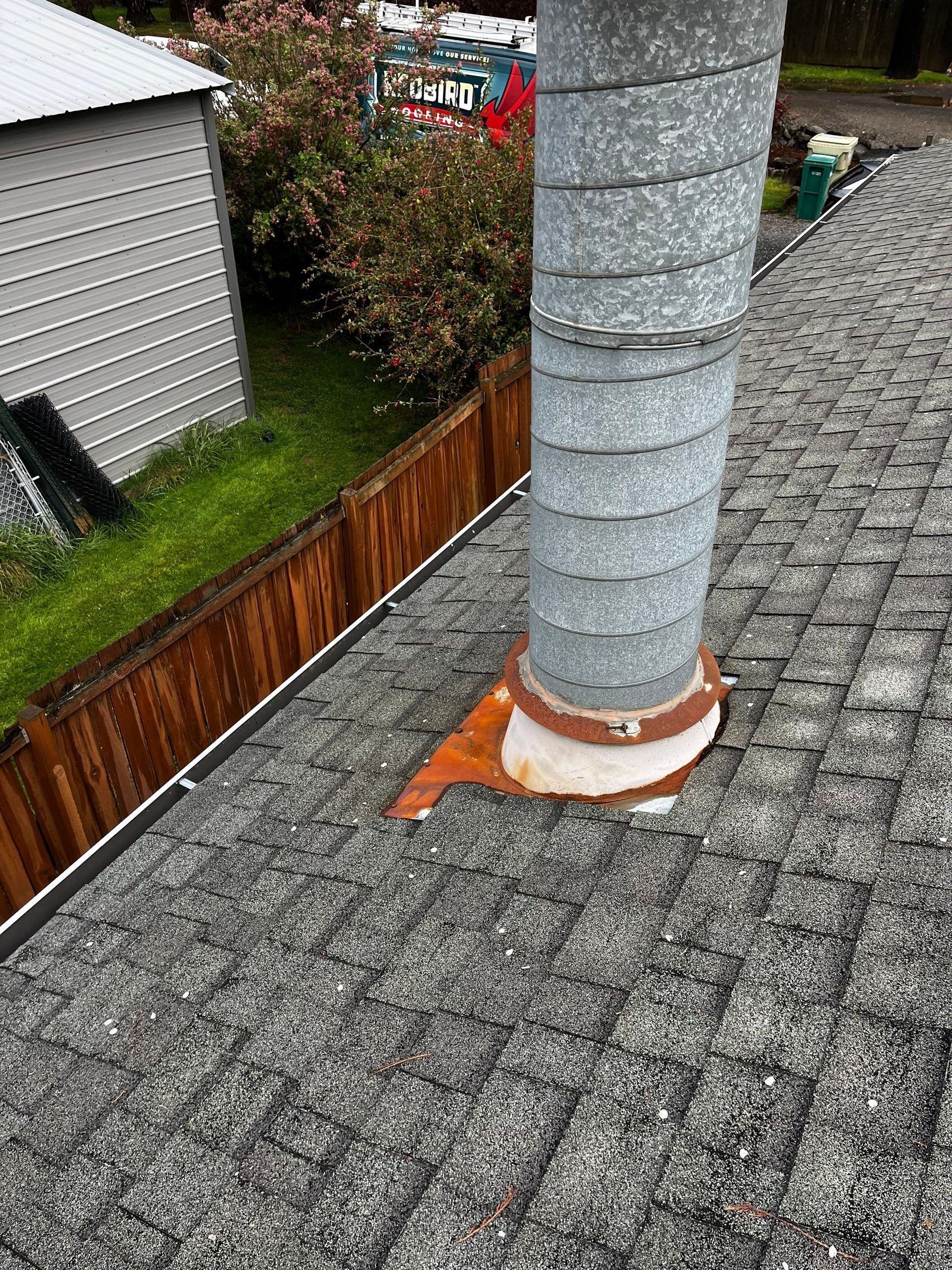 Gray chimney vent on a shingled roof beside a wooden fence and neighboring house