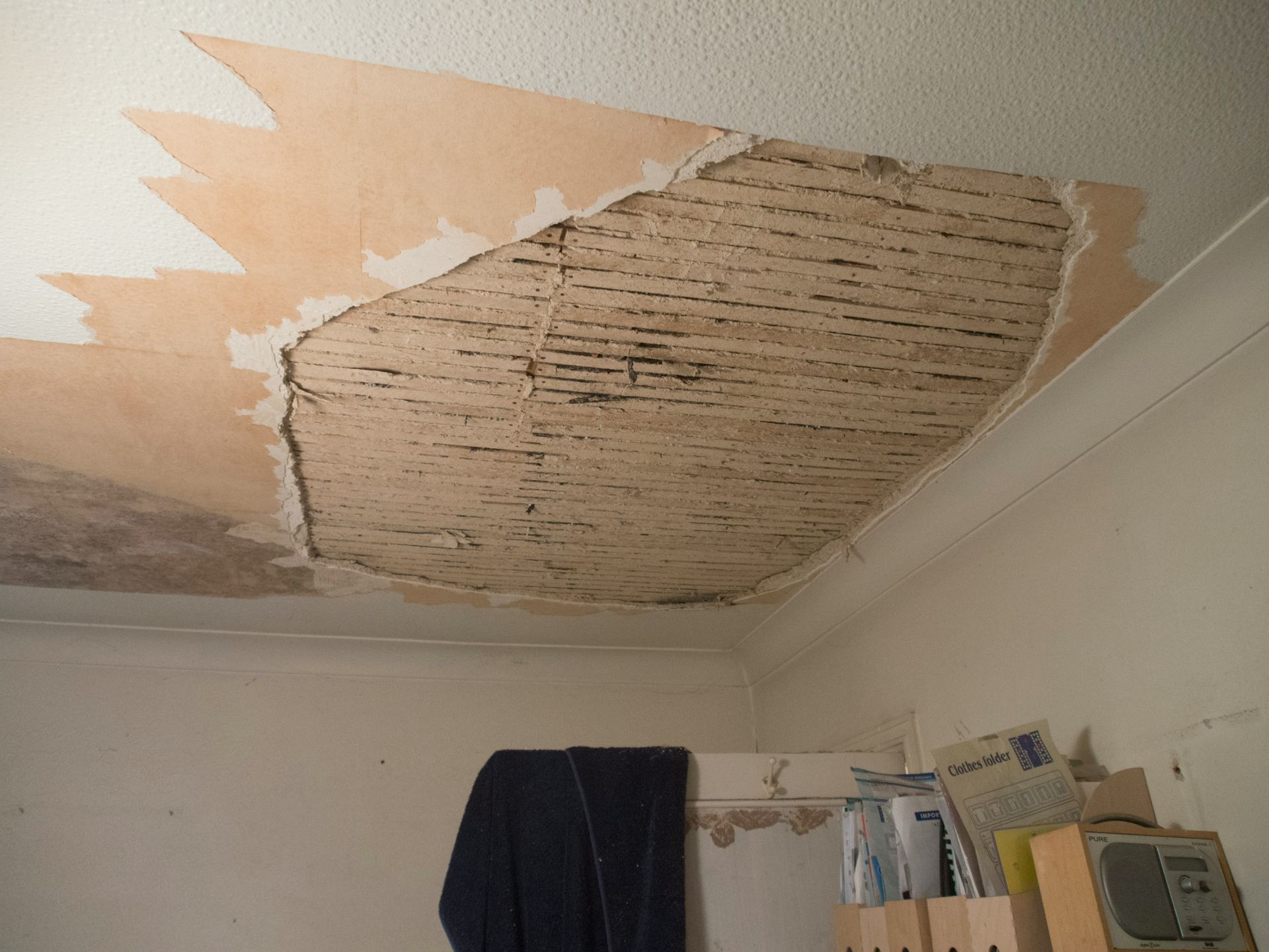 A damaged indoor ceiling with peeling plaster and exposed wooden lath strips.