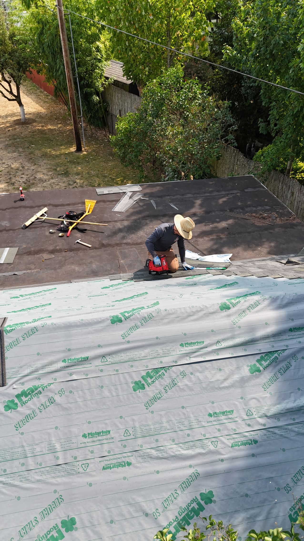 A person wearing a sun hat works on a residential roof, installing gray underlayment over a section of dark shingles.