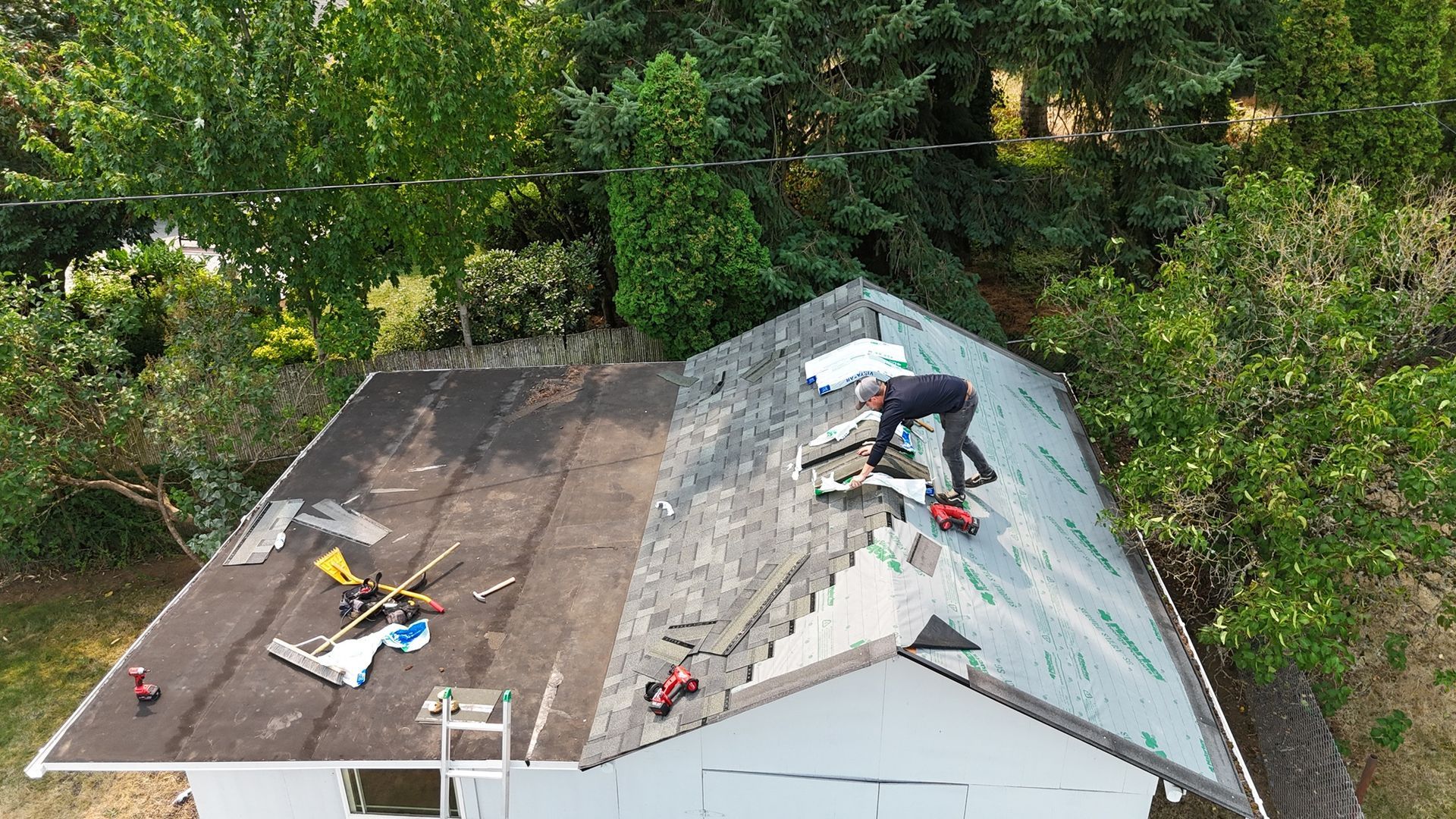 A worker is replacing shingles on a house roof surrounded by green trees.