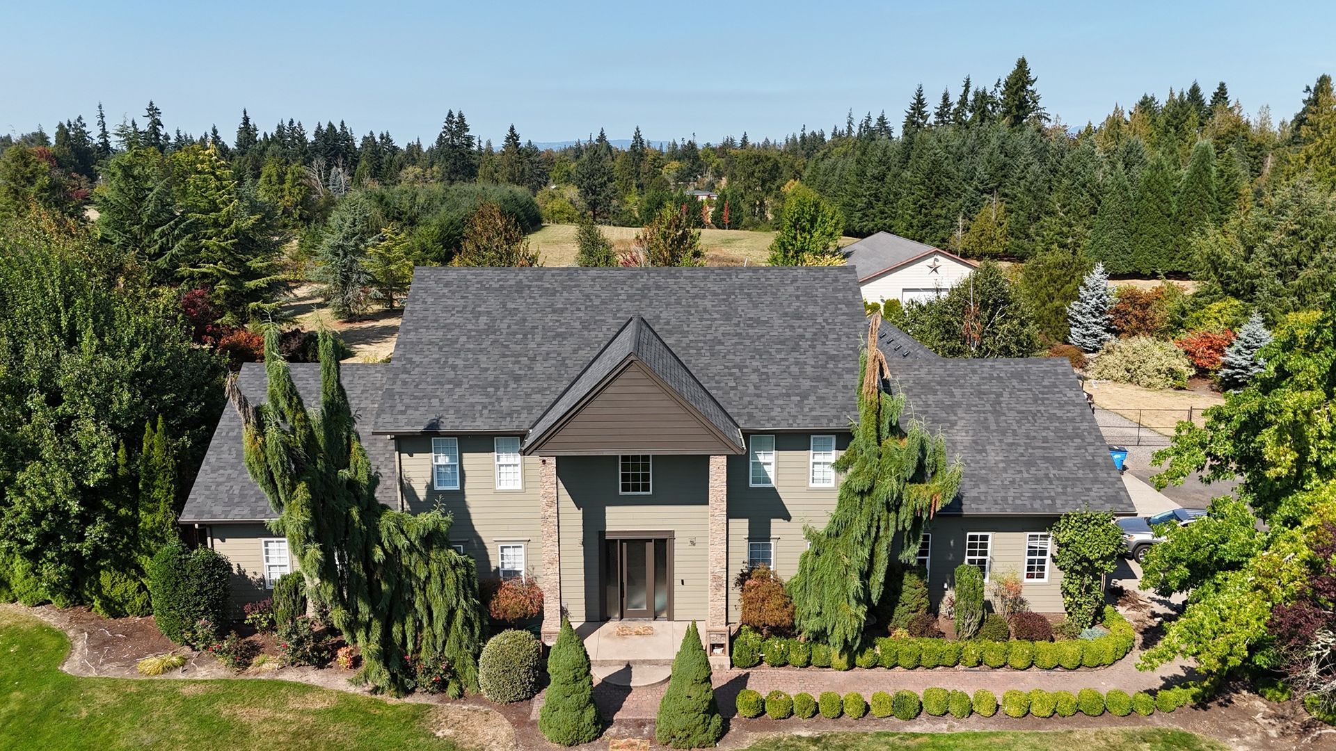 An aerial view of a two-story gray house with a dark shingled roof, surrounded by lush trees and greenery.