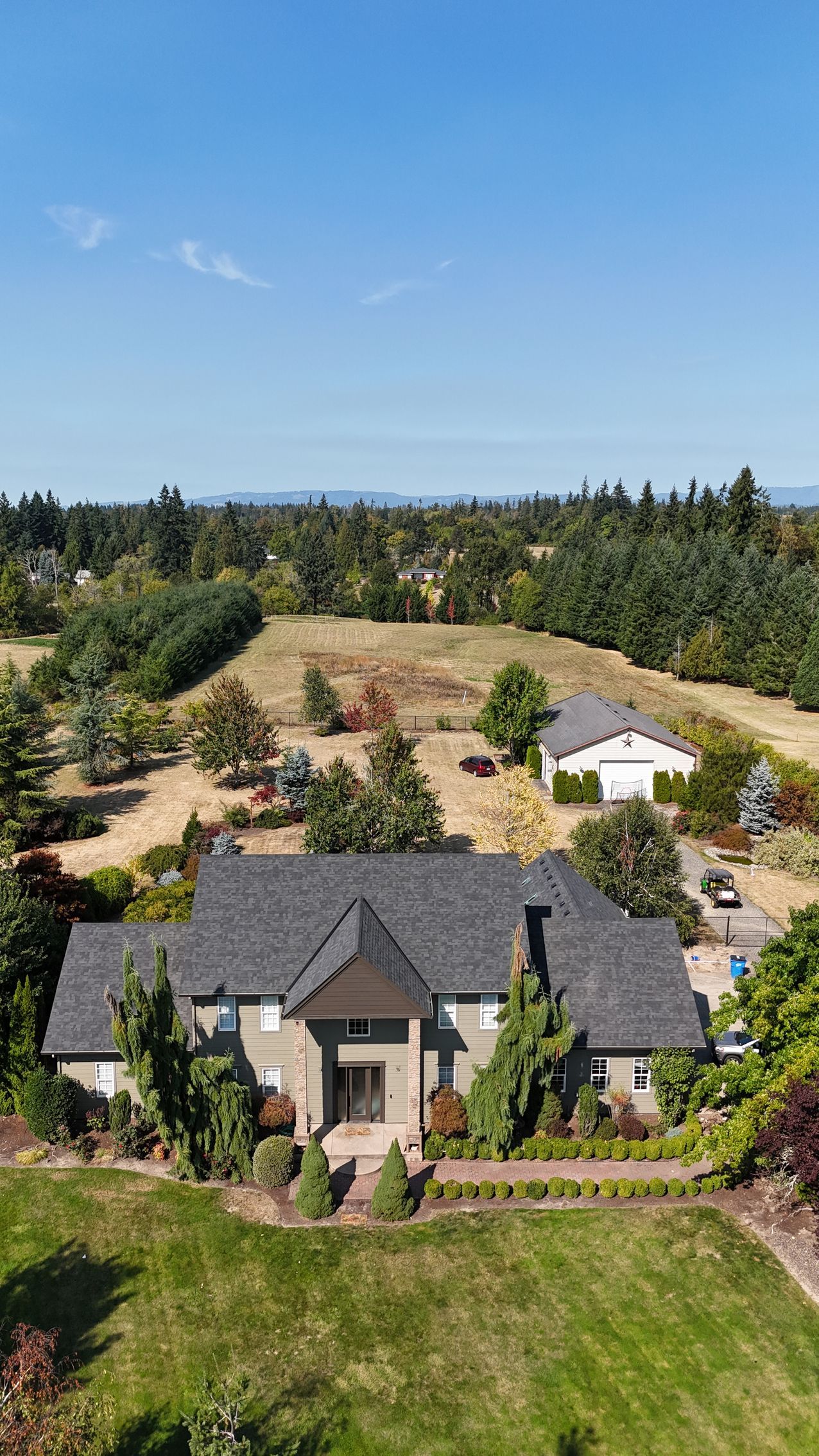 An aerial view of a large, gray-roofed house with a detached garage on a spacious property surrounded by trees.