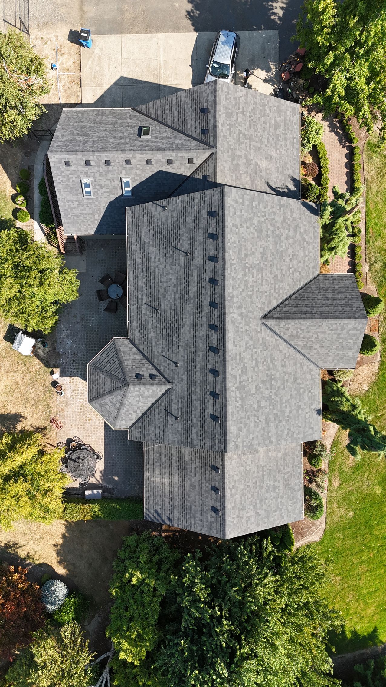 A top-down aerial view of a gray shingled roof on a residential home, surrounded by trees and a paved driveway.