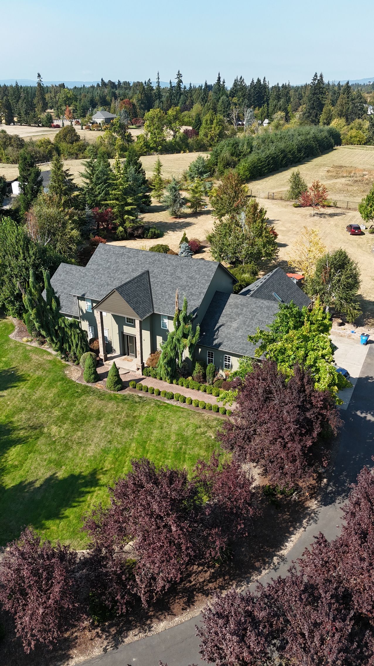 An aerial view of a large, gray-roofed house surrounded by green lawns, trees, and a paved driveway on a sunny day.