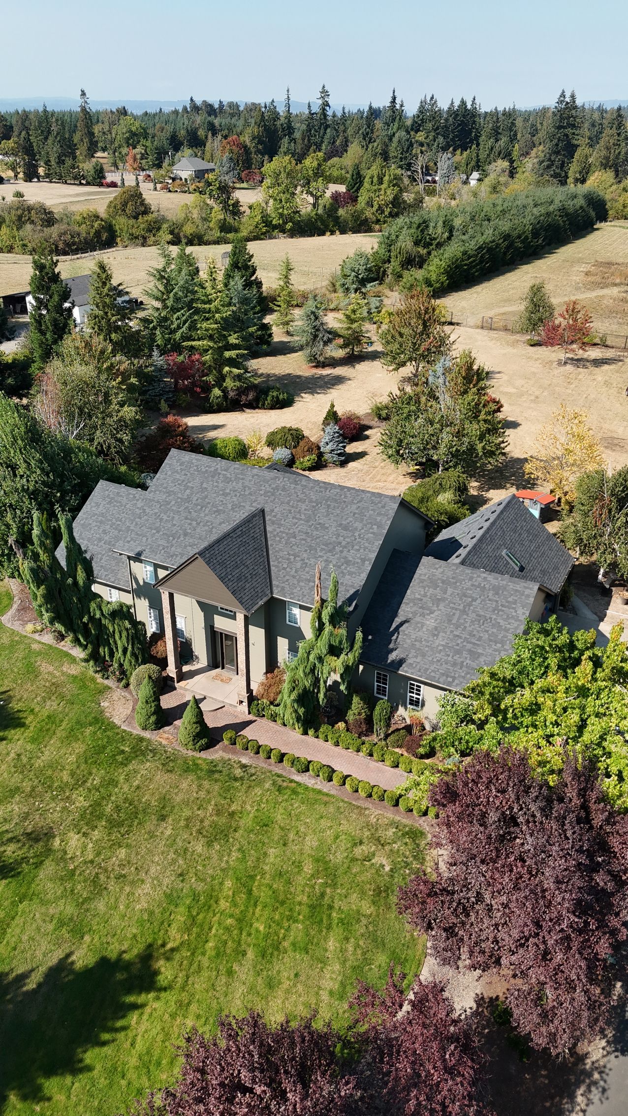 An aerial view of a large, gray-roofed house nestled in a lush, green landscape with surrounding trees and meadows.