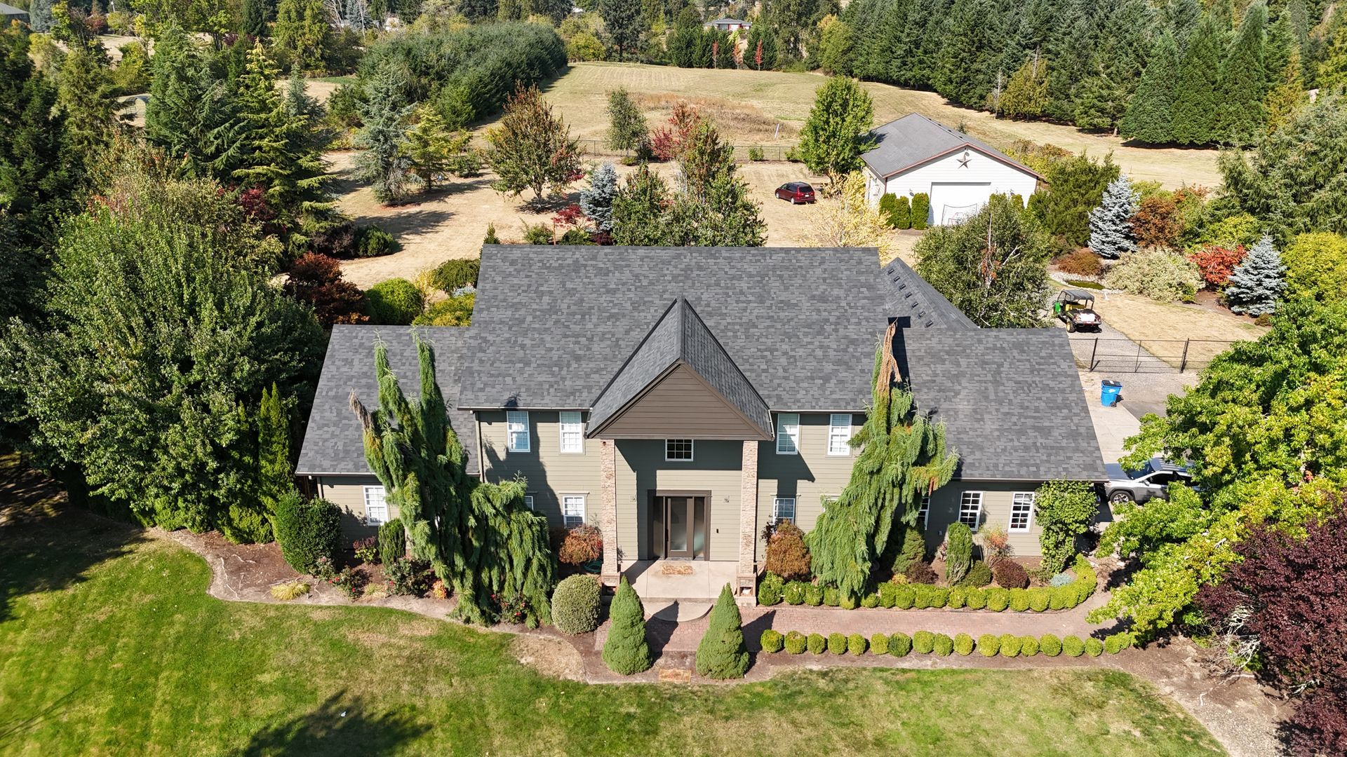 An aerial view of a gray-roofed, two-story suburban home surrounded by lush green trees, a grassy lawn, and a small garage.