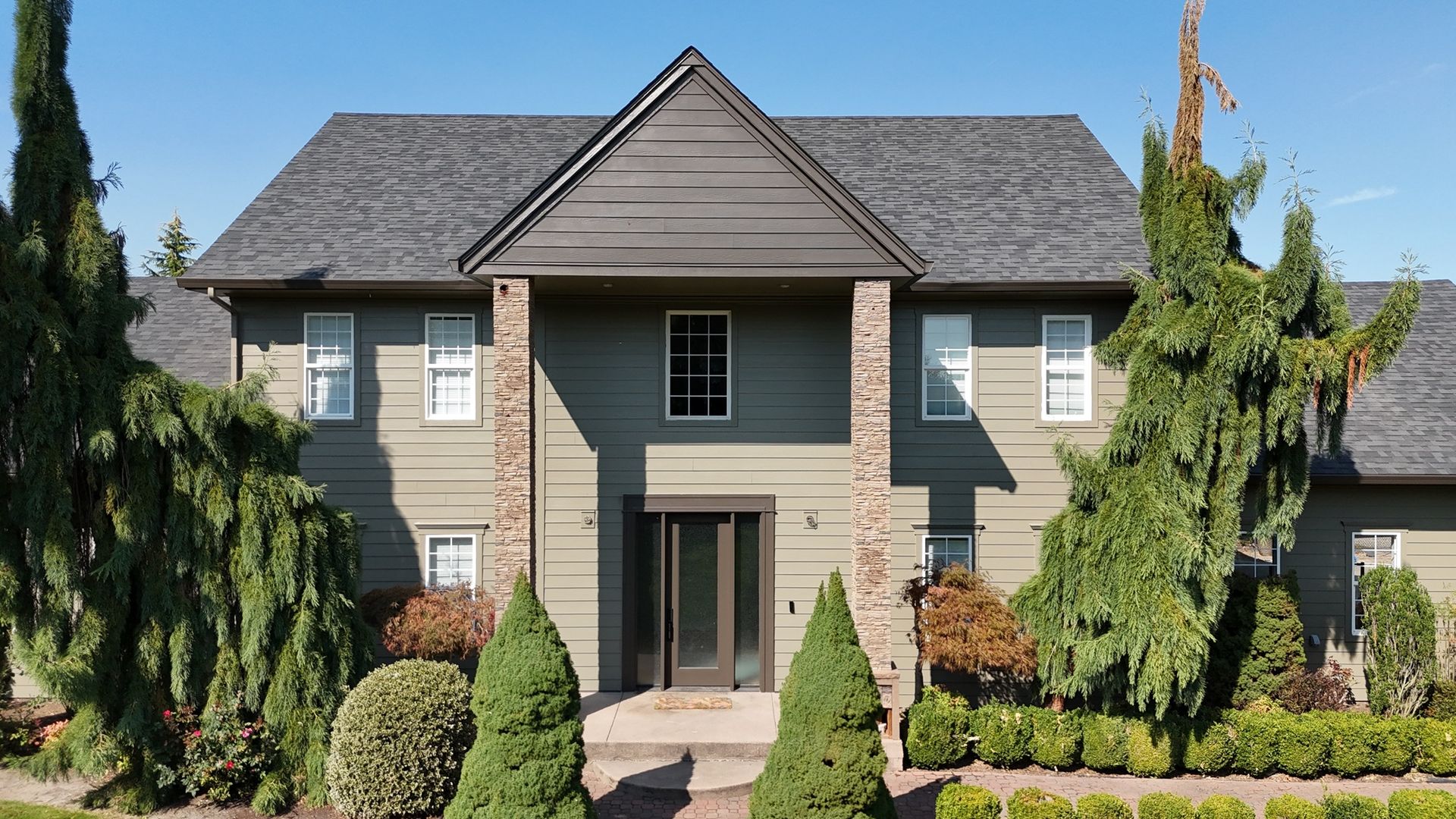 A two-story green house with a dark gabled roof, stone accents, and manicured landscaping under a clear blue sky.