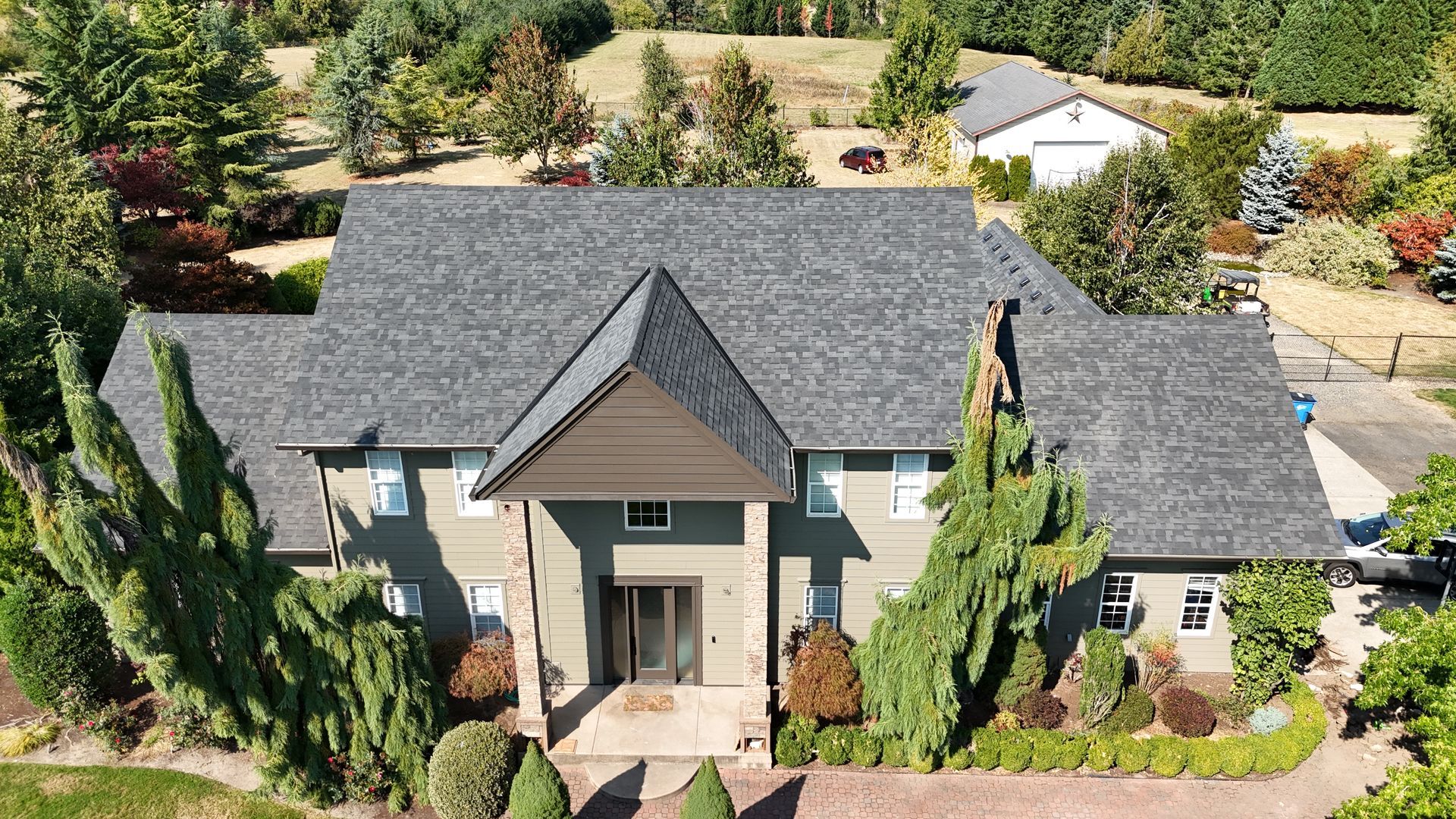An aerial view of a two-story gray house with a peaked roof and a stone entrance surrounded by trees and landscaping.