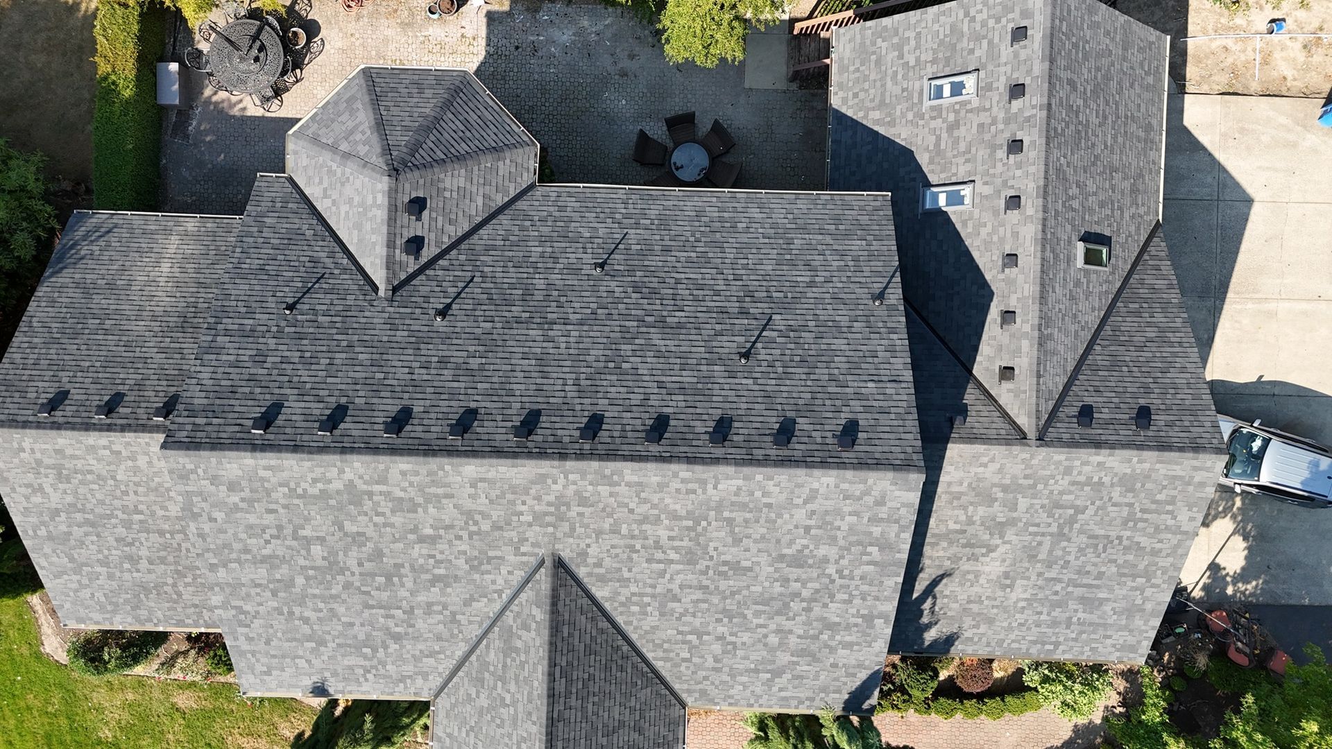 An aerial view of a dark grey shingled residential roof with multiple peaks, valleys, and skylights.