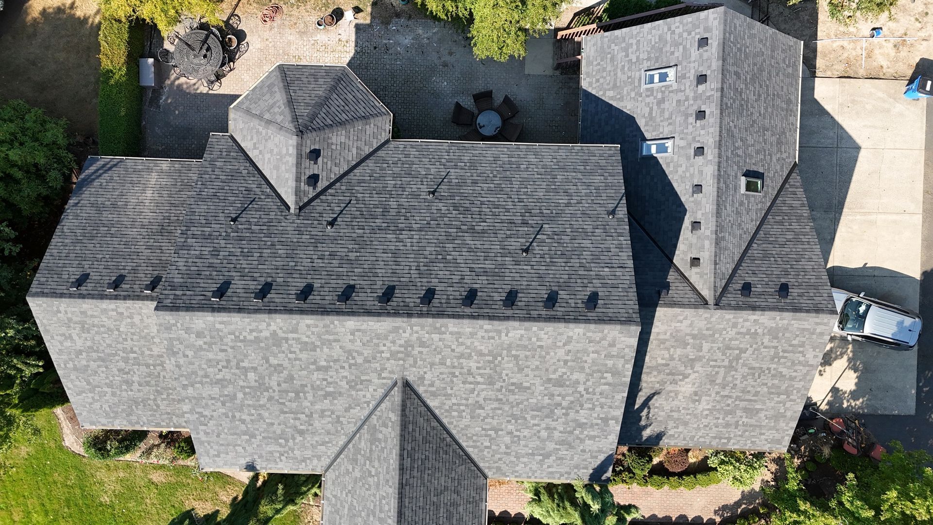 Aerial view of a complex, multi-gabled gray shingle roof on a residential home surrounded by trees and a paved driveway.