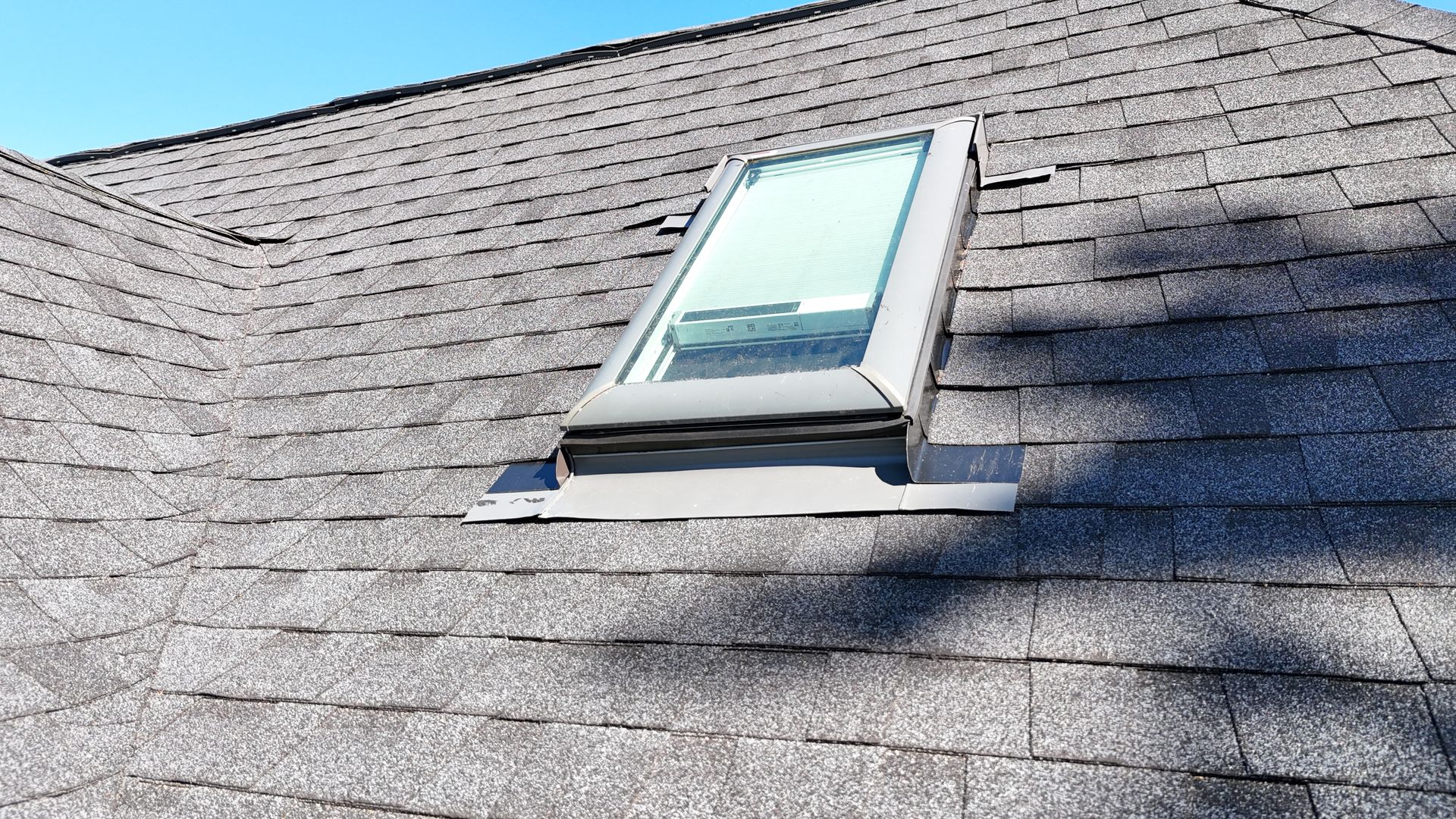 A rectangular skylight installed on a gray asphalt shingle roof against a clear blue sky.