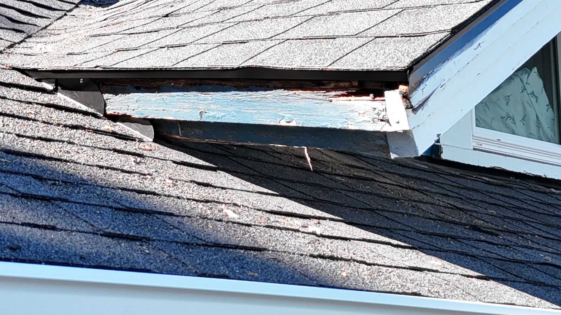 A section of a residential roof showing a damaged, rotting wooden trim board underneath the shingled roof overhang.