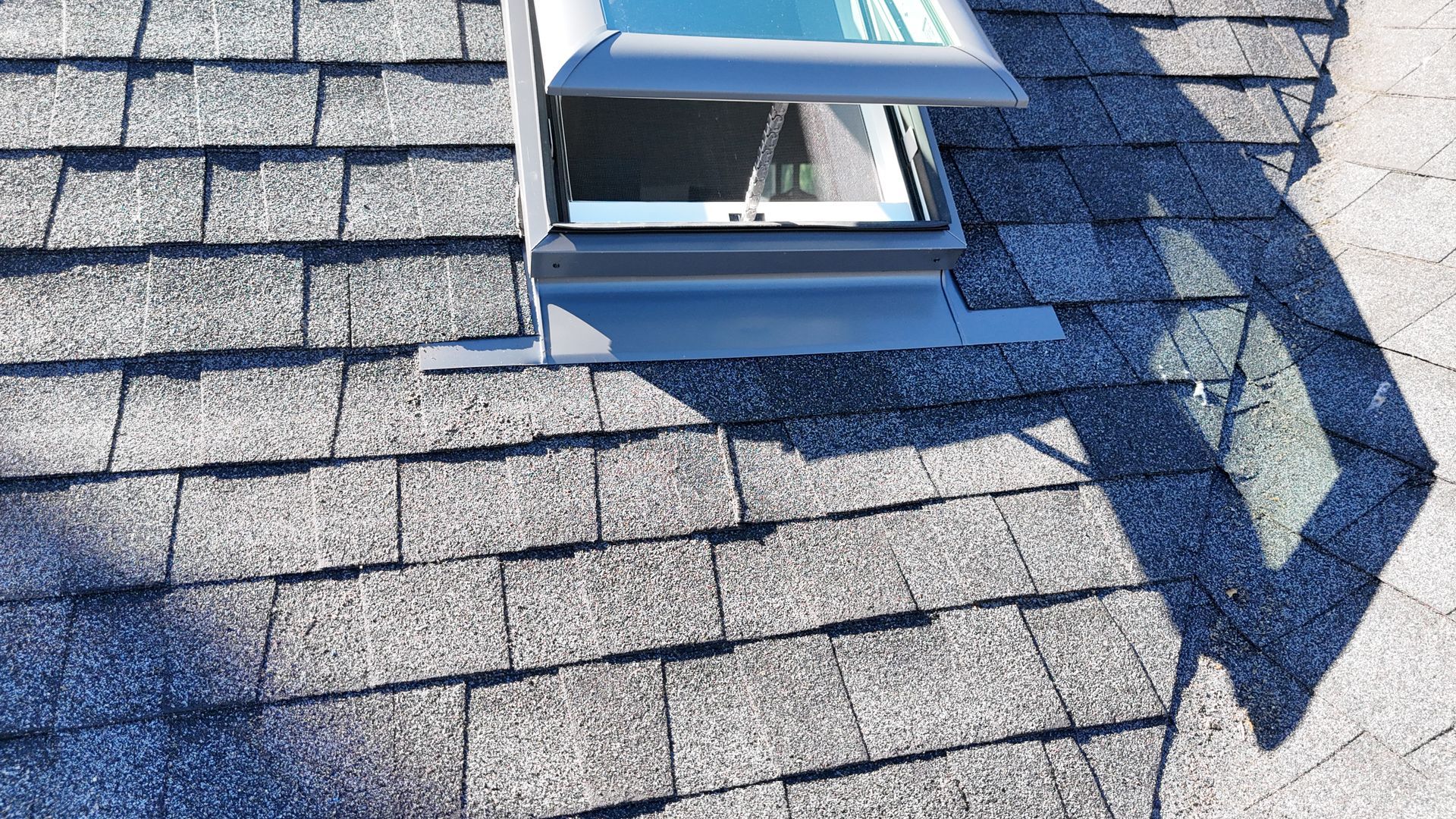 An open skylight installed on a dark gray asphalt shingle roof.