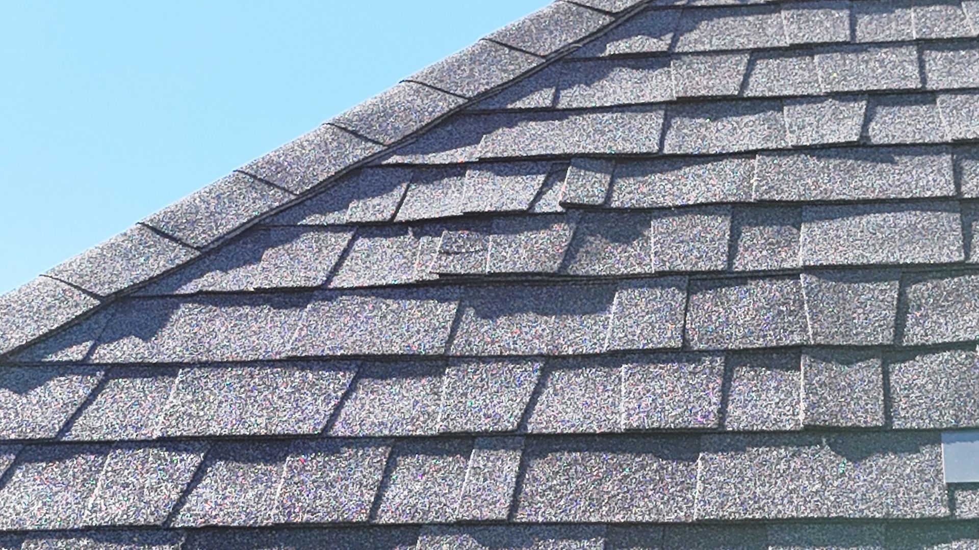 A close-up view of grey, textured asphalt shingles on a pitched residential roof against a clear blue sky.