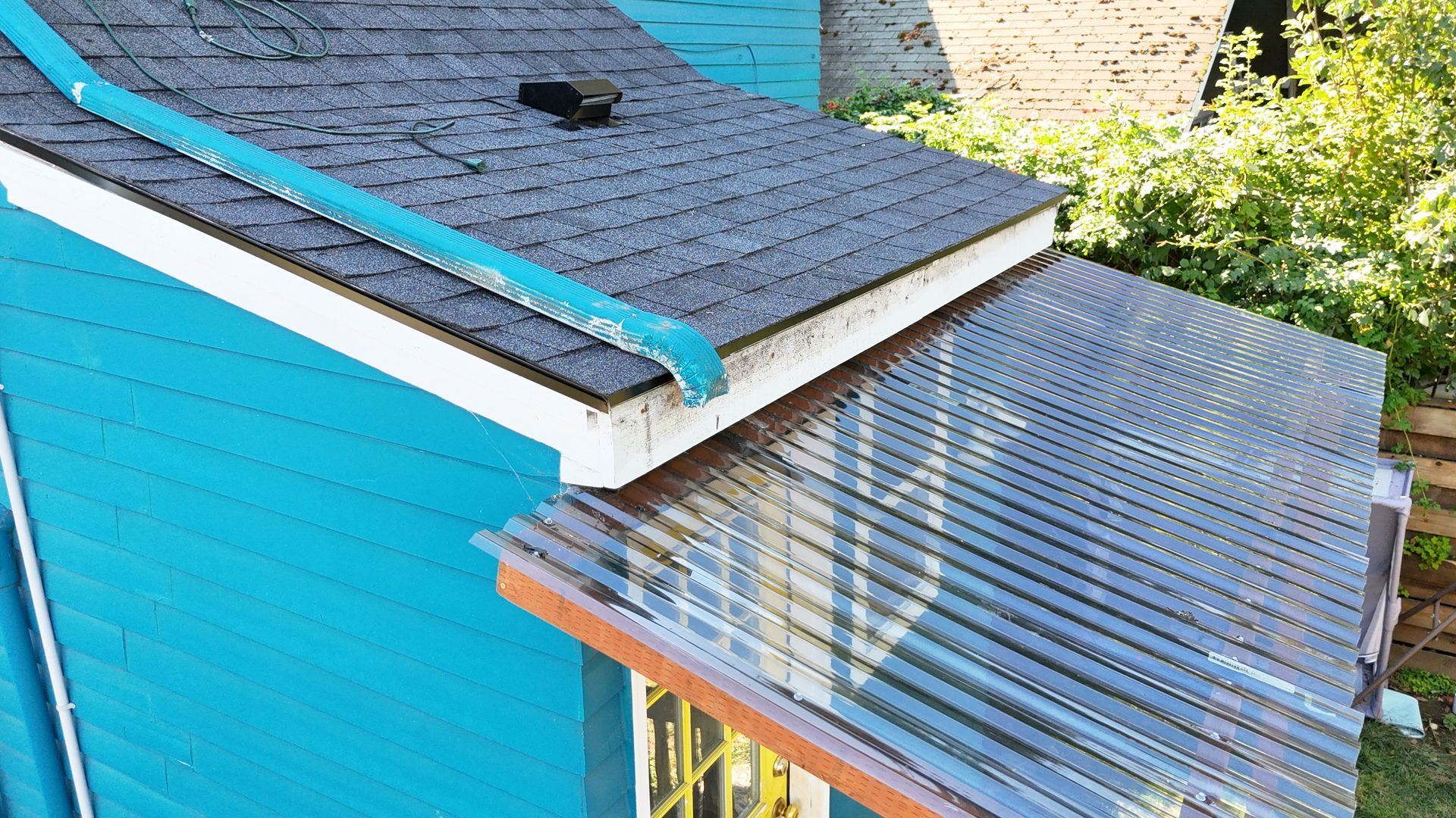 A teal-colored house exterior featuring a grey shingled roof with a blue trim and an attached, clear corrugated awning.