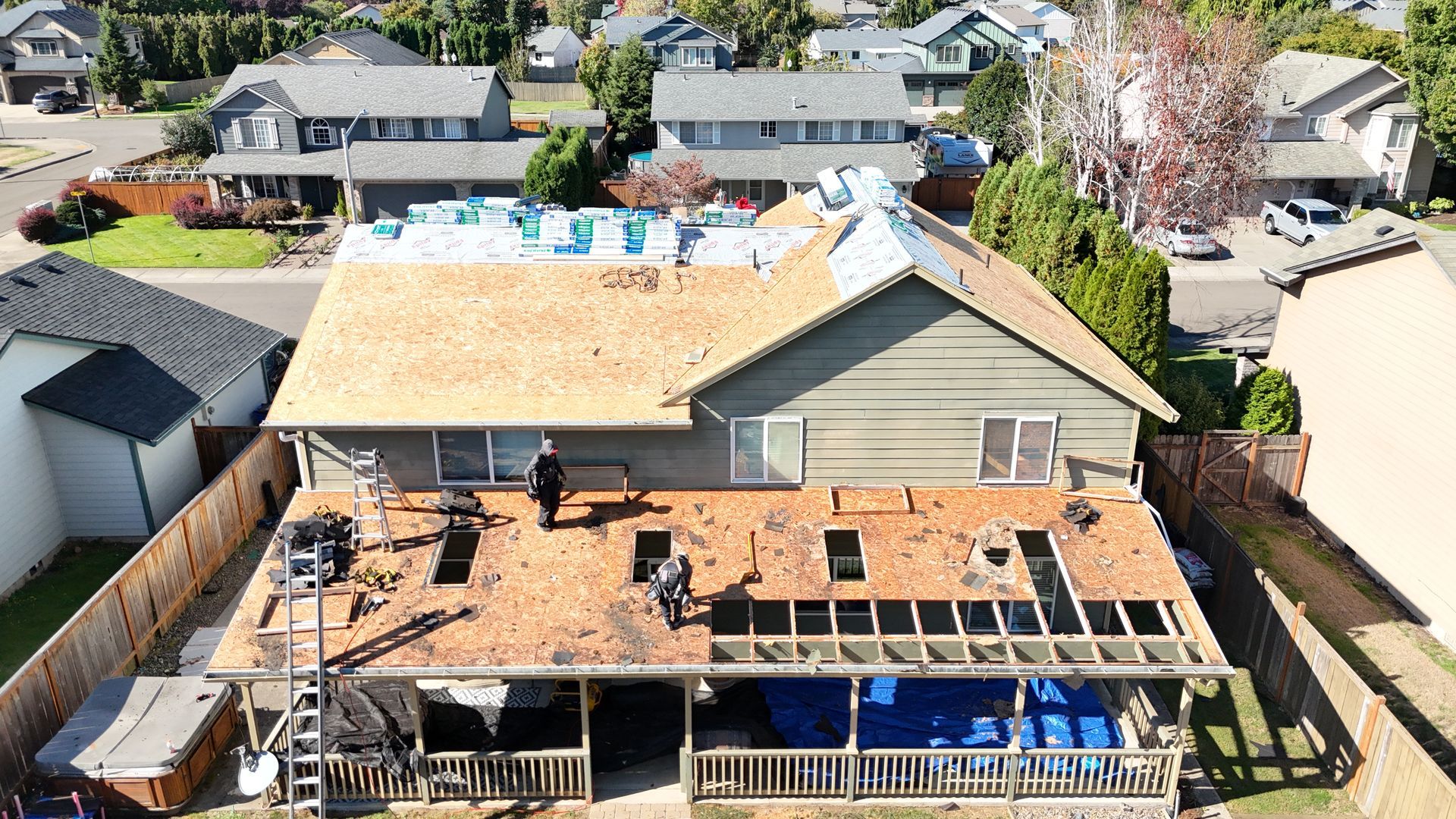 Aerial view of workers replacing the roof of a suburban house, showing plywood decking and roof construction in progress.