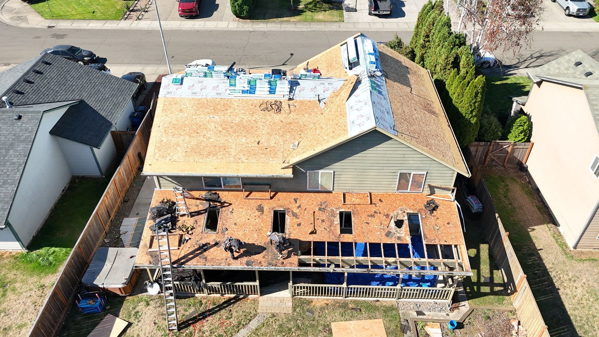 An aerial view of a house undergoing roof renovations, showing exposed wooden decking, construction materials, and workers.