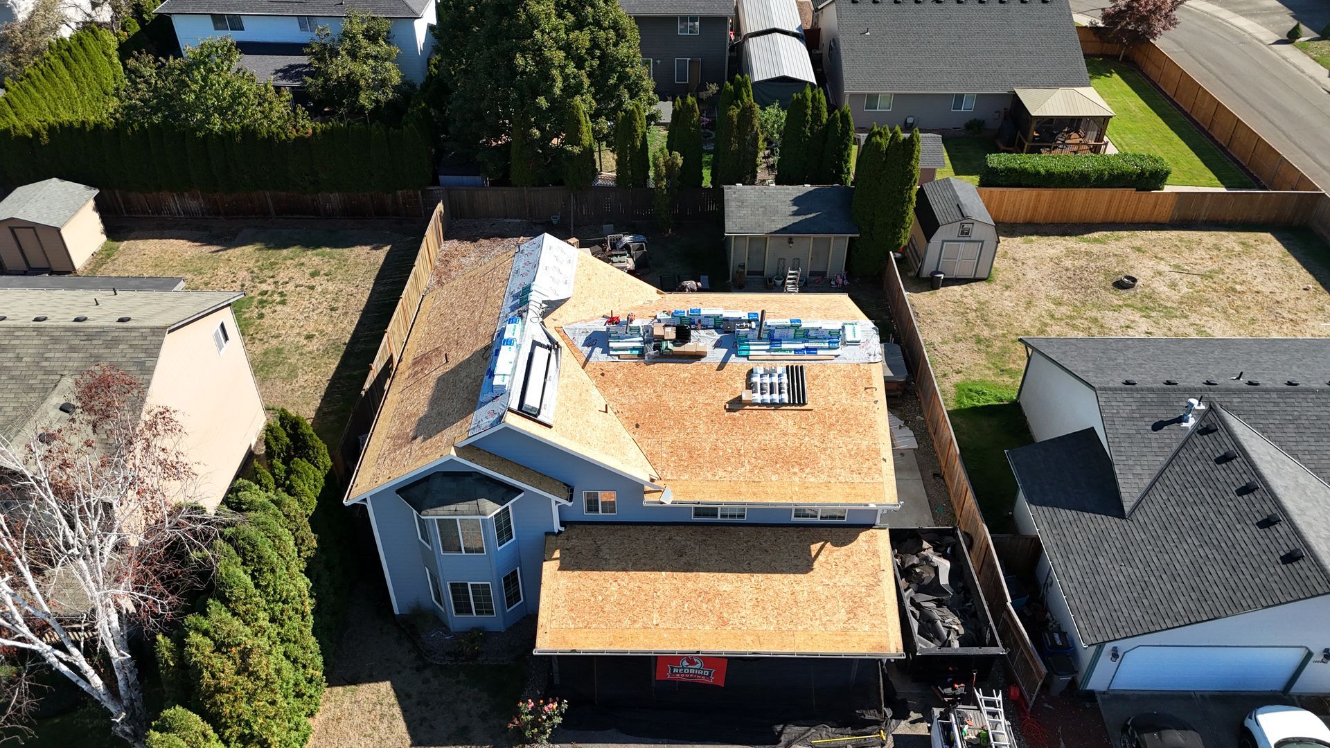 An aerial view of a house undergoing roof replacement, featuring exposed plywood sheathing and roofing materials.