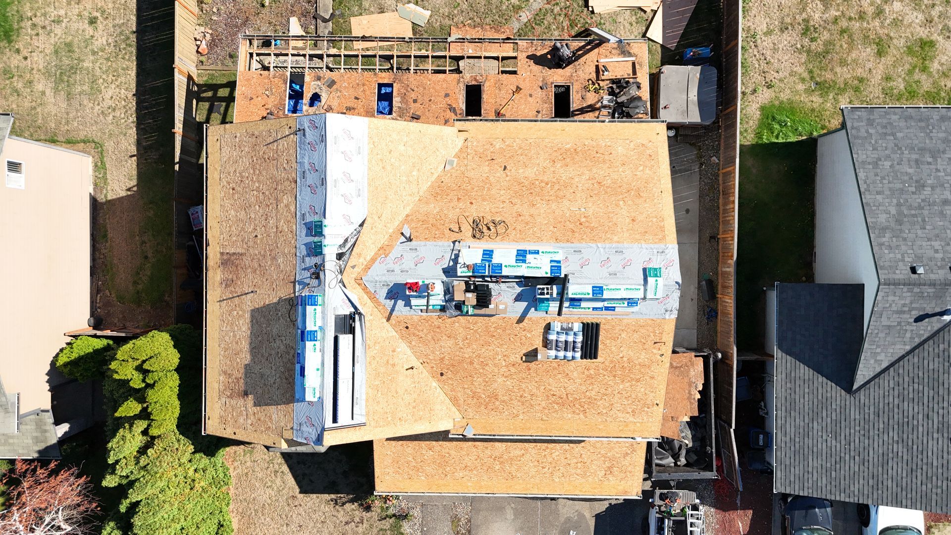 A high-angle aerial view of a house roof under construction, showing plywood sheathing and rolls of underlayment.