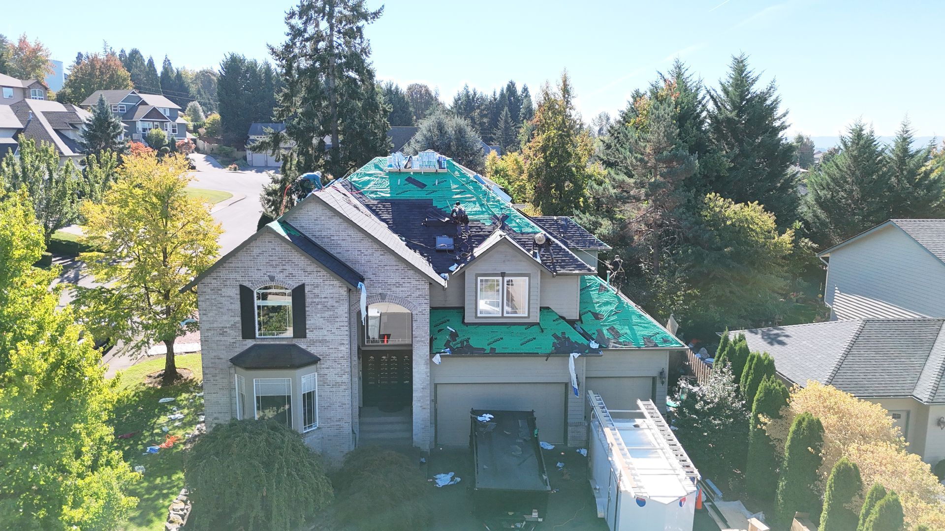 An aerial view of a suburban home undergoing roof repairs, with sections of the roof covered in green underlayment.