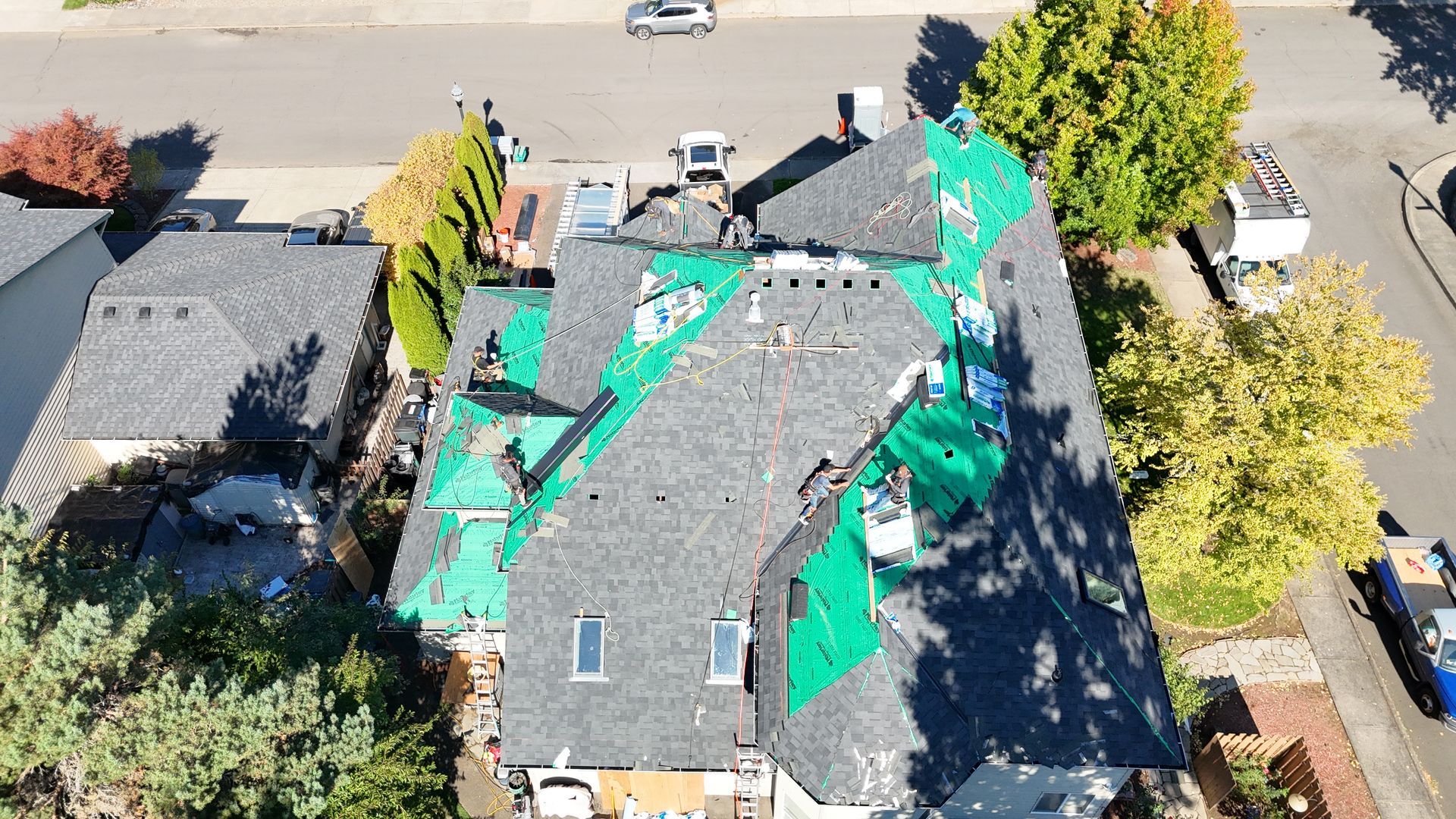 An aerial view of a residential roof undergoing construction, featuring grey shingles and bright green underlayment.