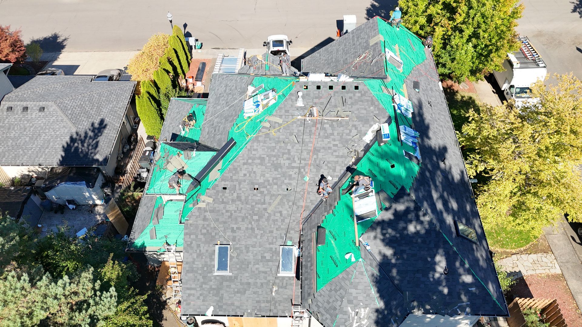 An aerial view of a house roof undergoing repairs, with sections of gray shingles removed, exposing green underlayment.