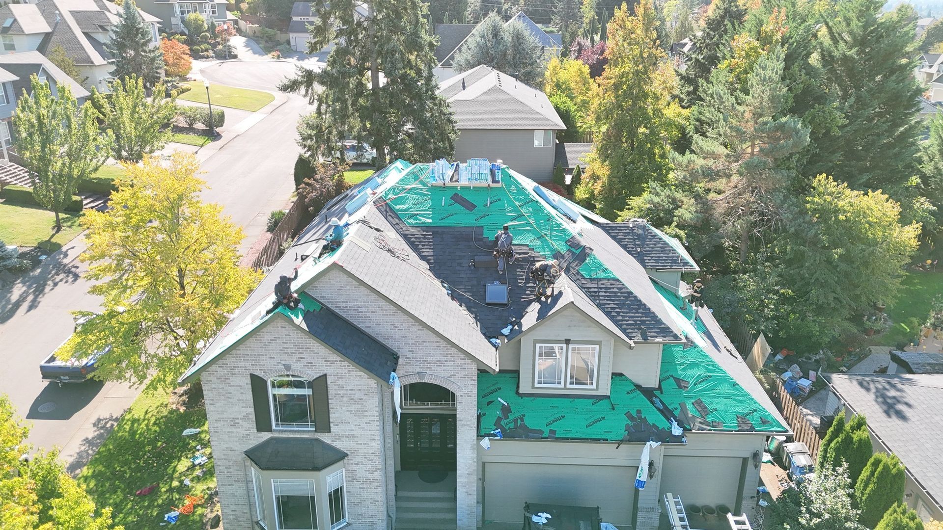 An aerial view shows a two-story house undergoing roof repairs, with sections of green underlayment exposed.