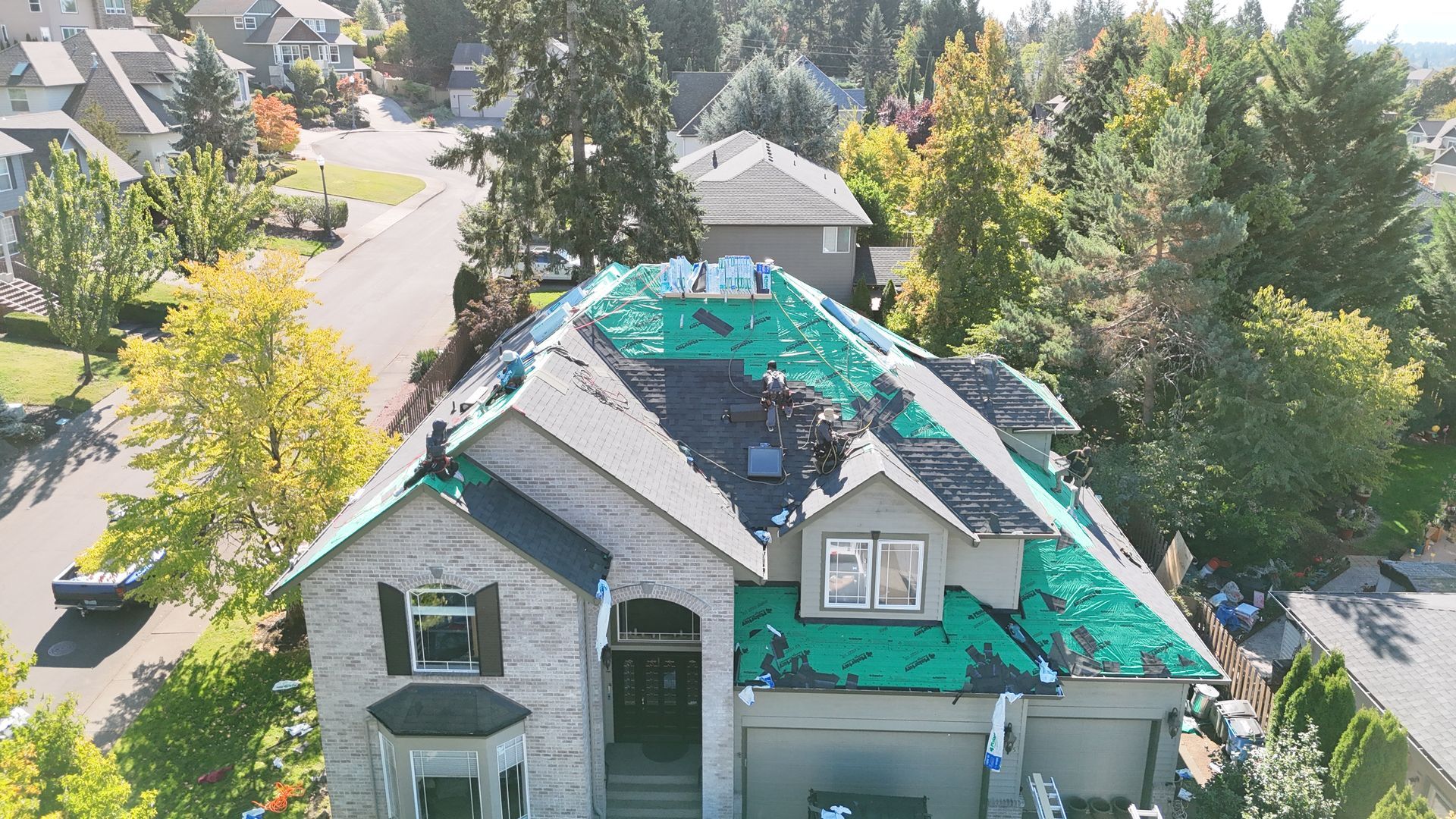 An aerial view of a suburban house during roof renovation, showing shingles removed to reveal green protective underlay.