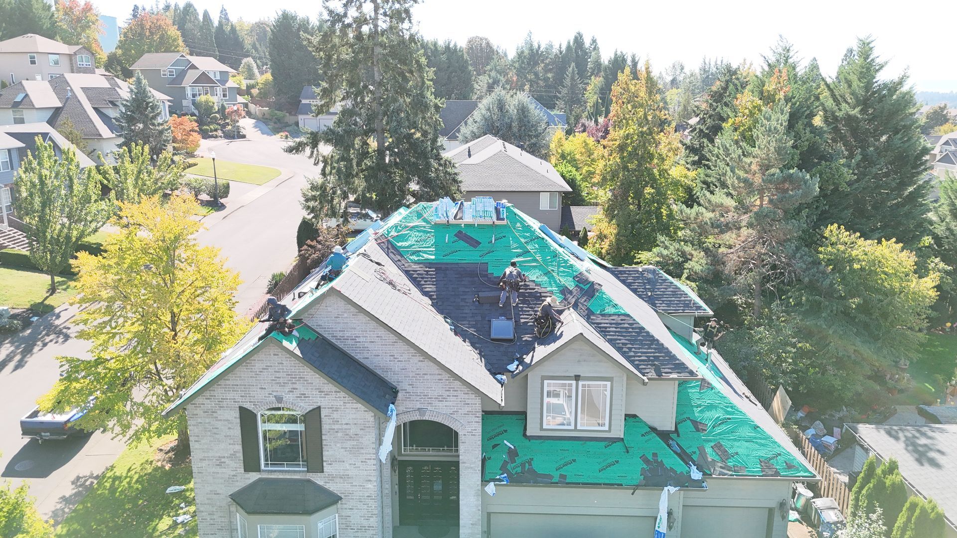 Aerial view of workers replacing shingles on a house roof with green underlayment exposed.