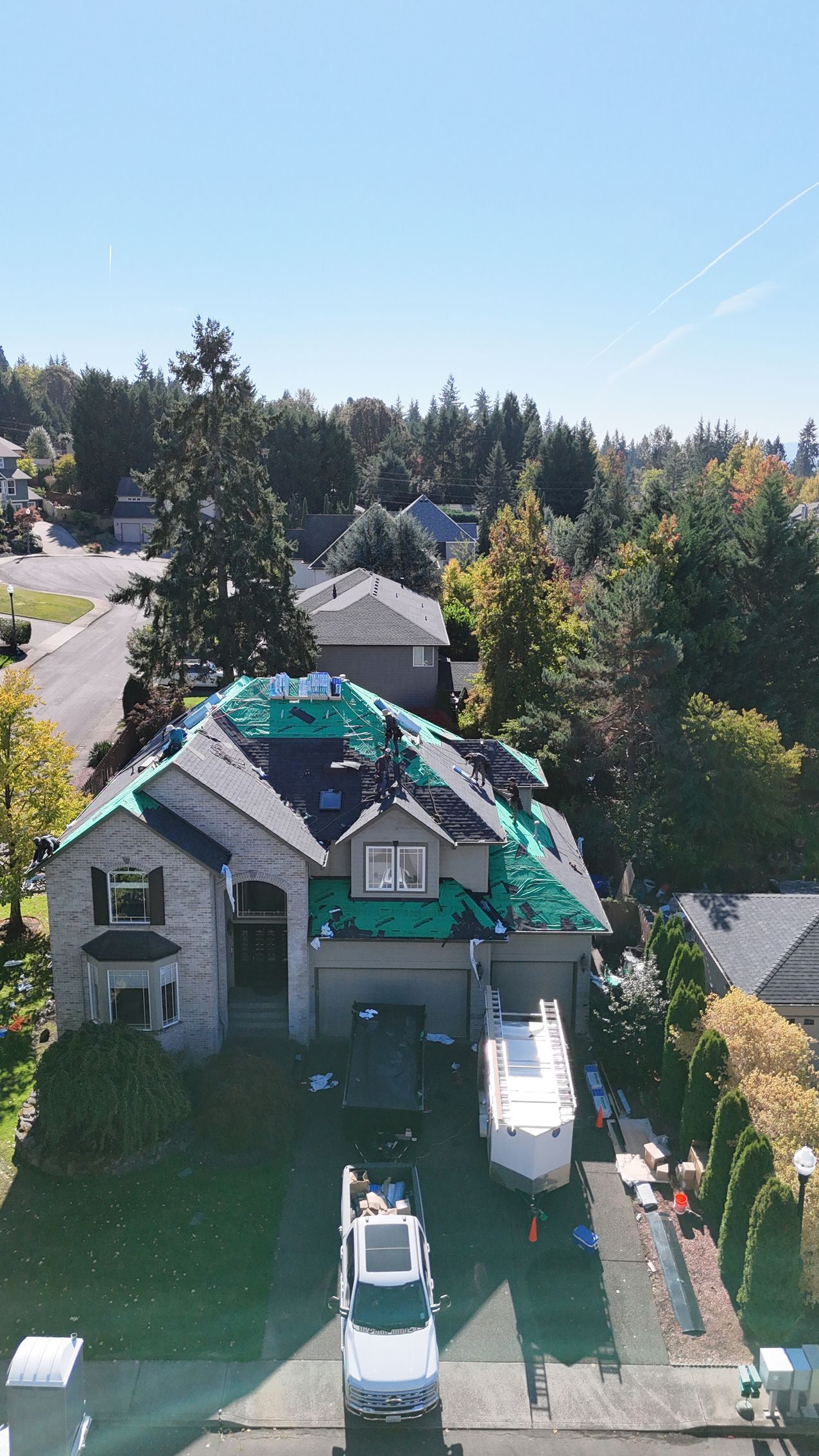 An aerial view of a two-story house under construction with a green tarp covering parts of its roof, and vehicles parked.