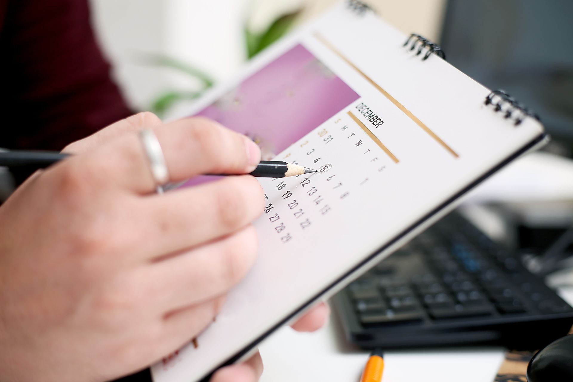 A person holding a pen points to a date on a desk calendar, with a computer keyboard visible in the blurred background.