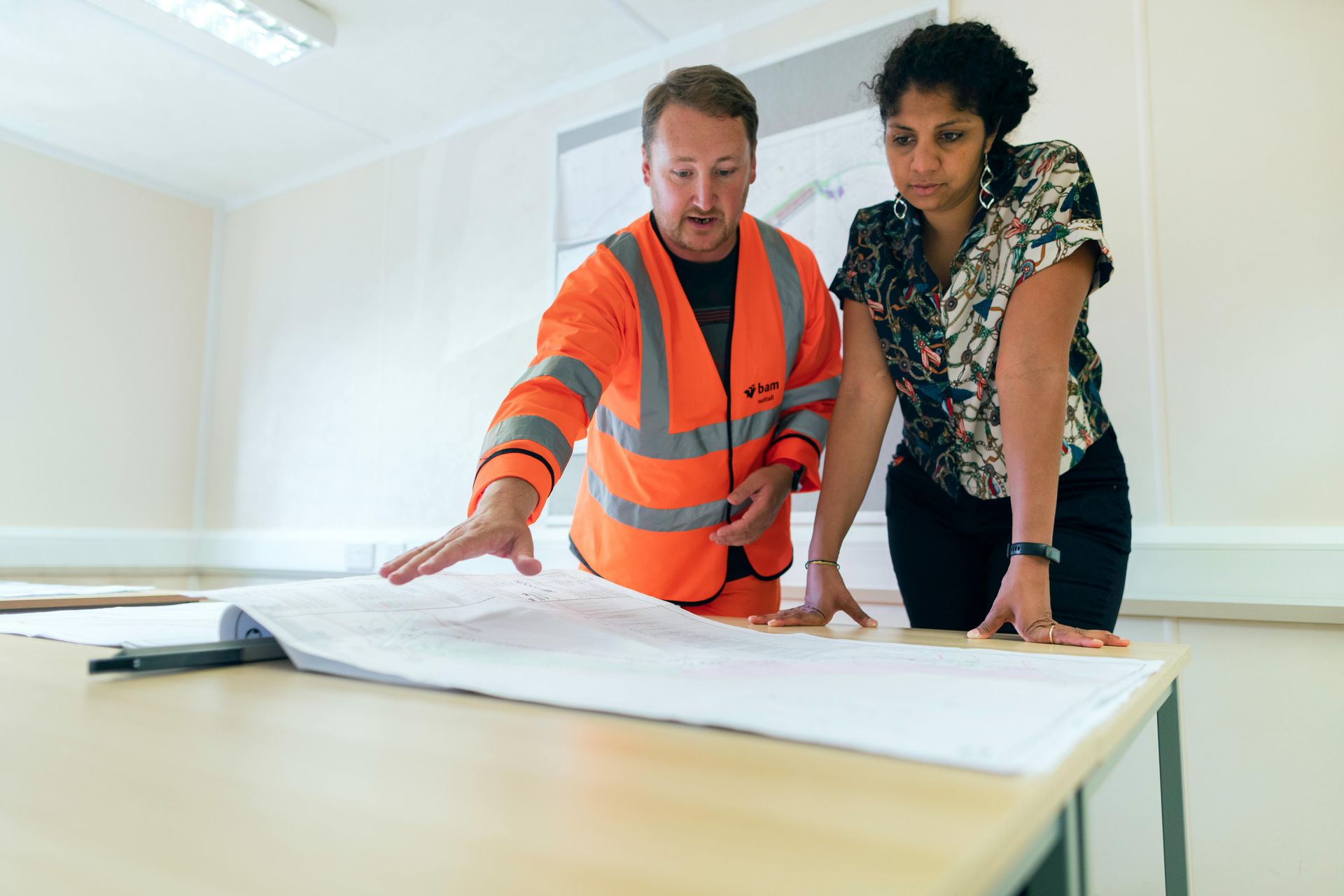 A person in an orange high-visibility vest and a person in a patterned blouse review a large blueprint at a desk.