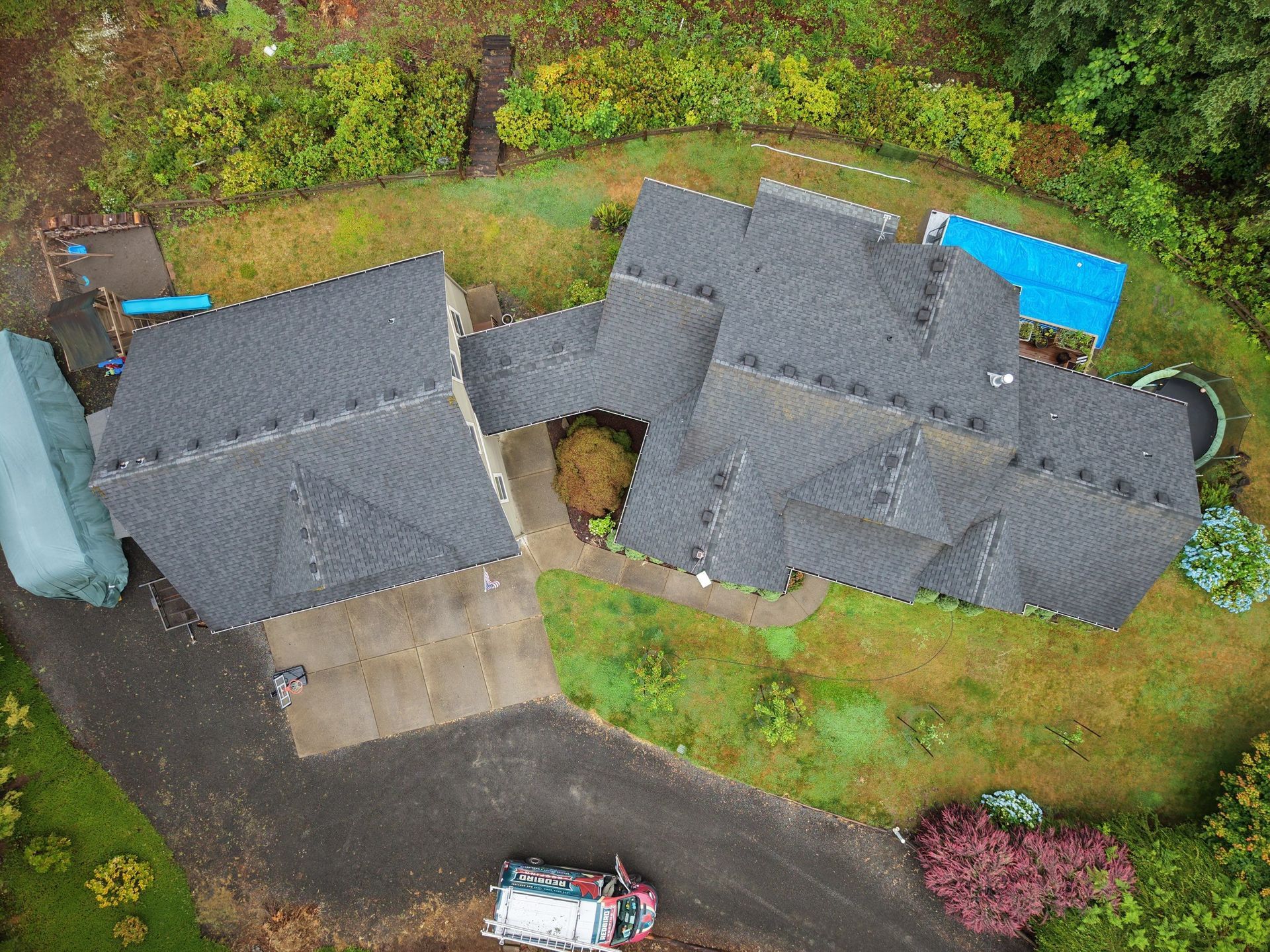 Aerial view of a house with dark gray roofs, driveway, and green yard bordered by trees