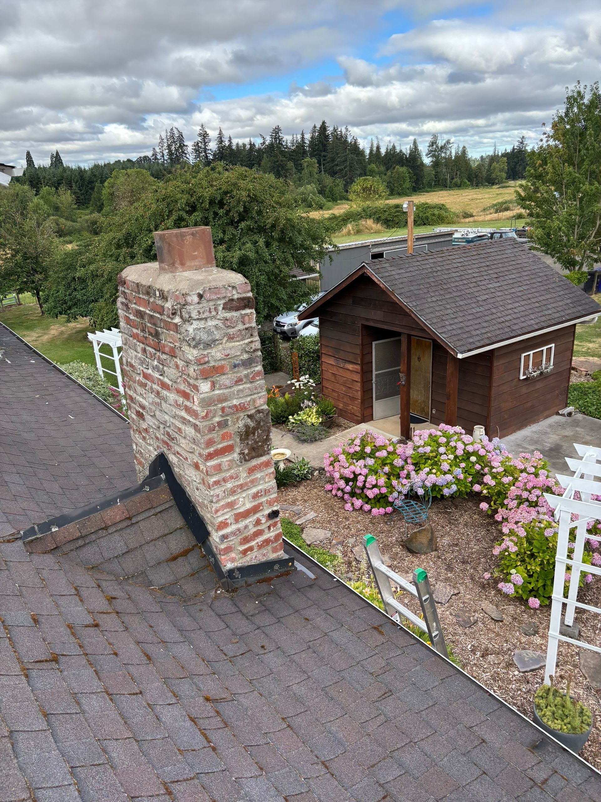 Miniature rustic house and brick chimney model in a garden, with flowers, gravel paths, and cloudy sky.