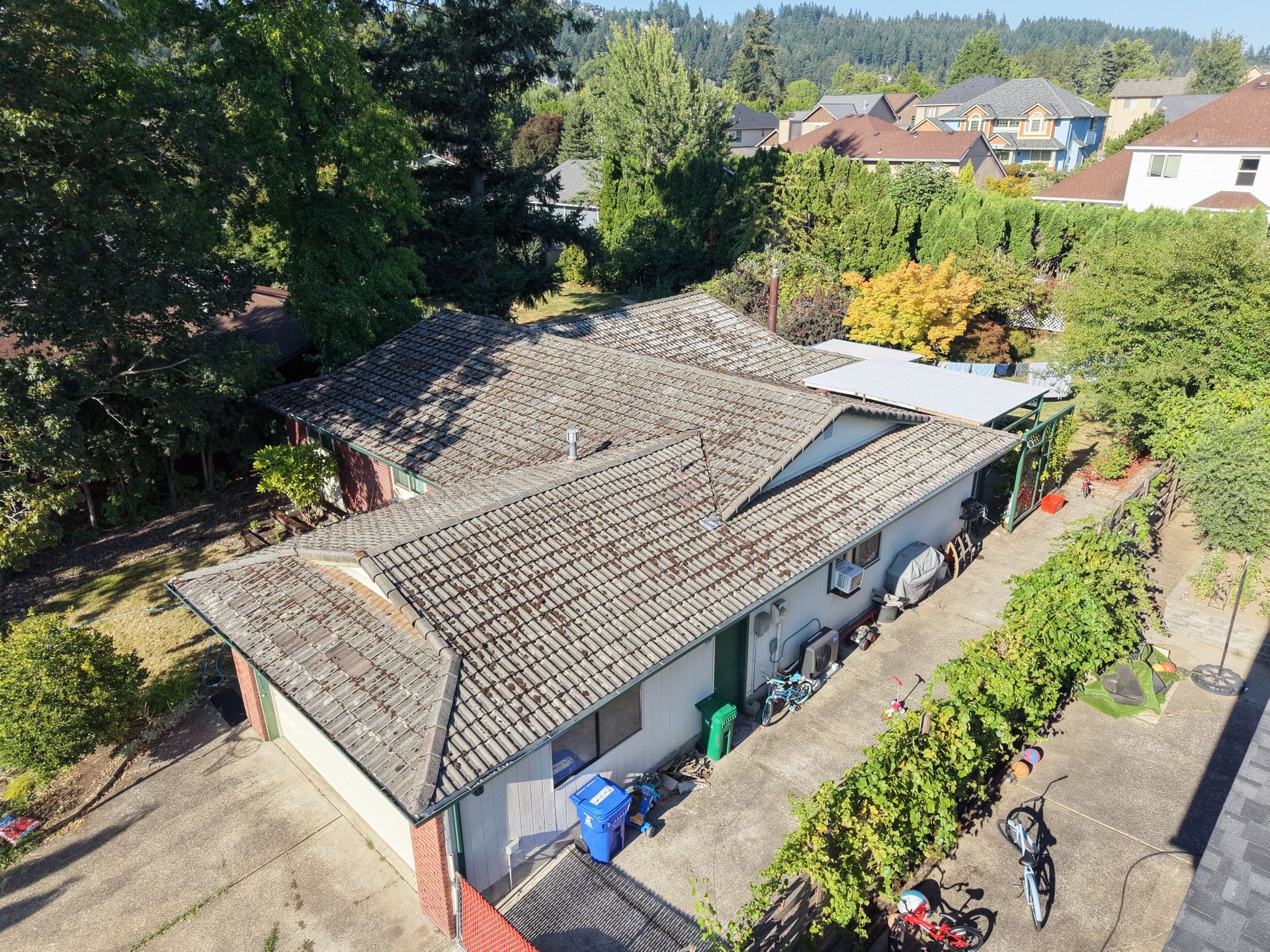 Aerial view of a long gray-roofed house with a skylight, surrounded by trees and neighboring homes.