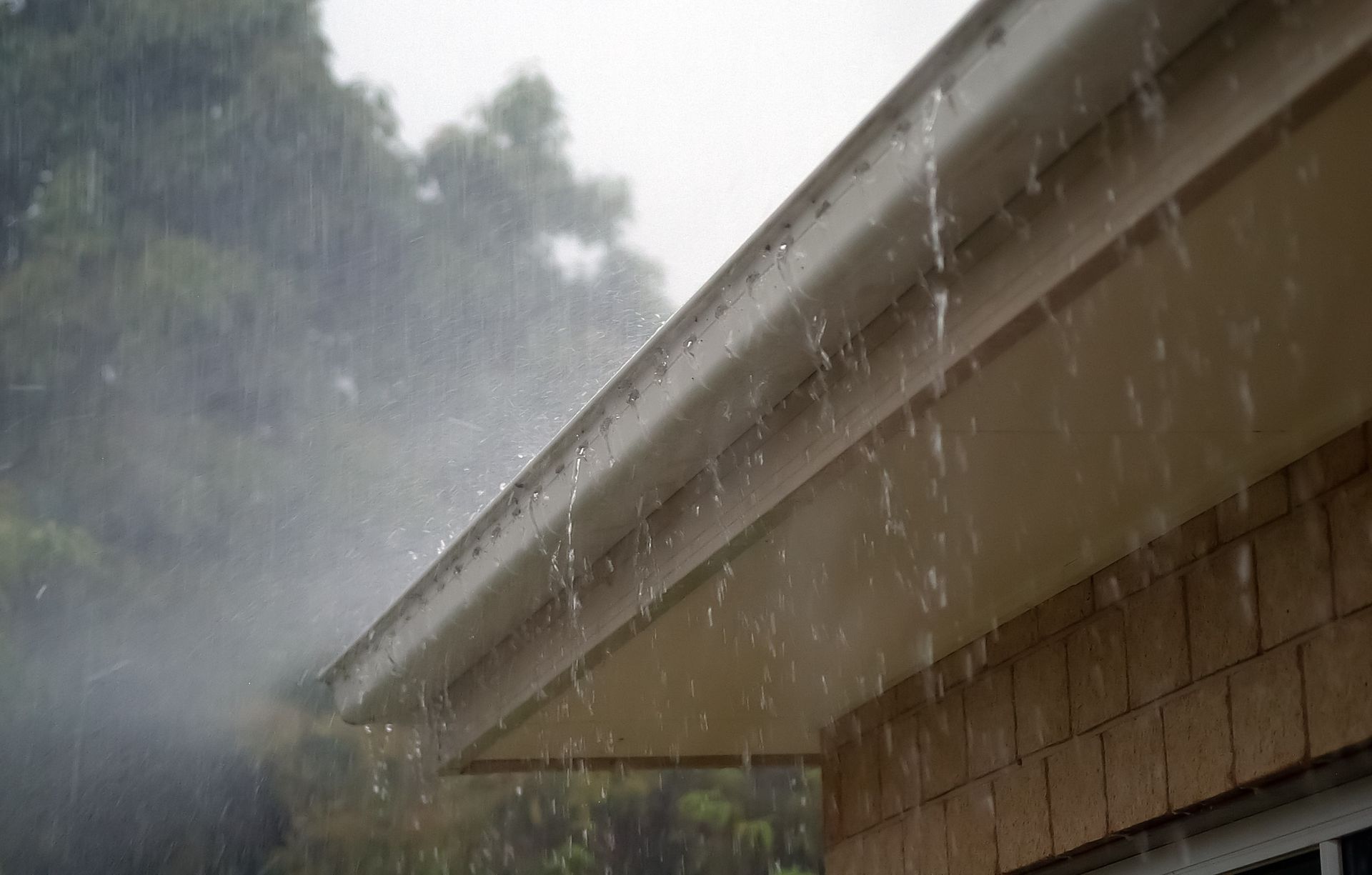 Heavy rain falling from the eaves of a building with light-colored siding and a brick wall.