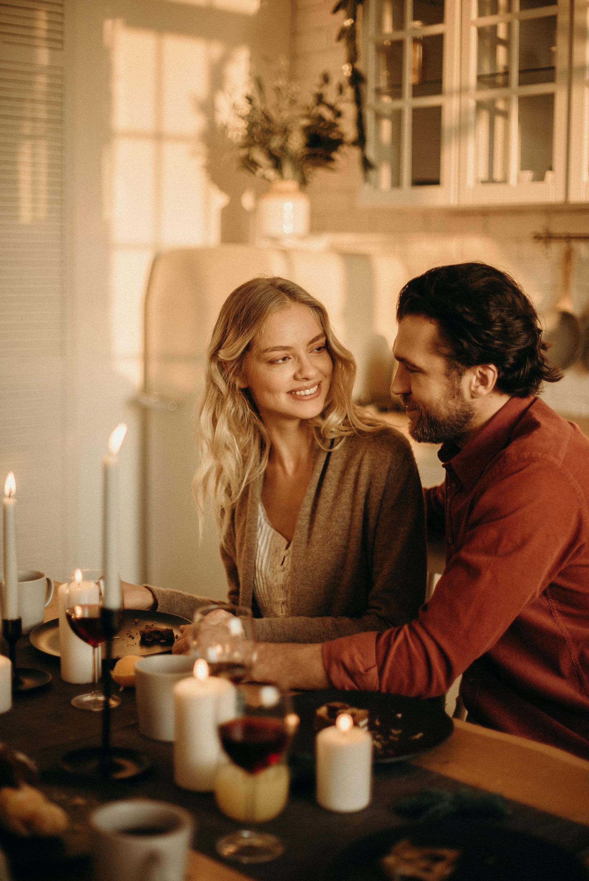 Couple at candlelit dinner, smiling and holding hands. Warm tones, kitchen setting.