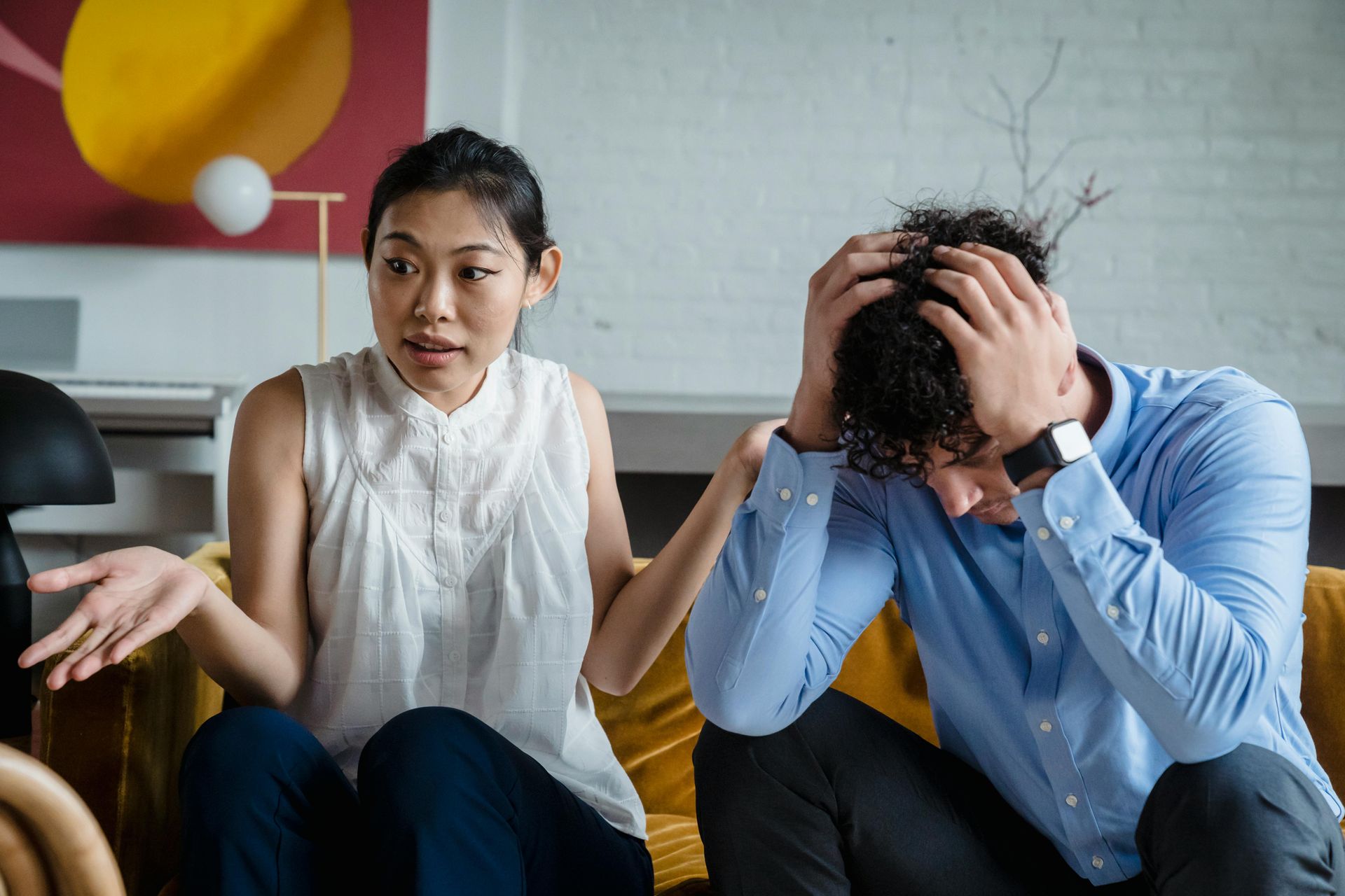 Woman gesturing and speaking, man with head in hands on a couch, indoors.