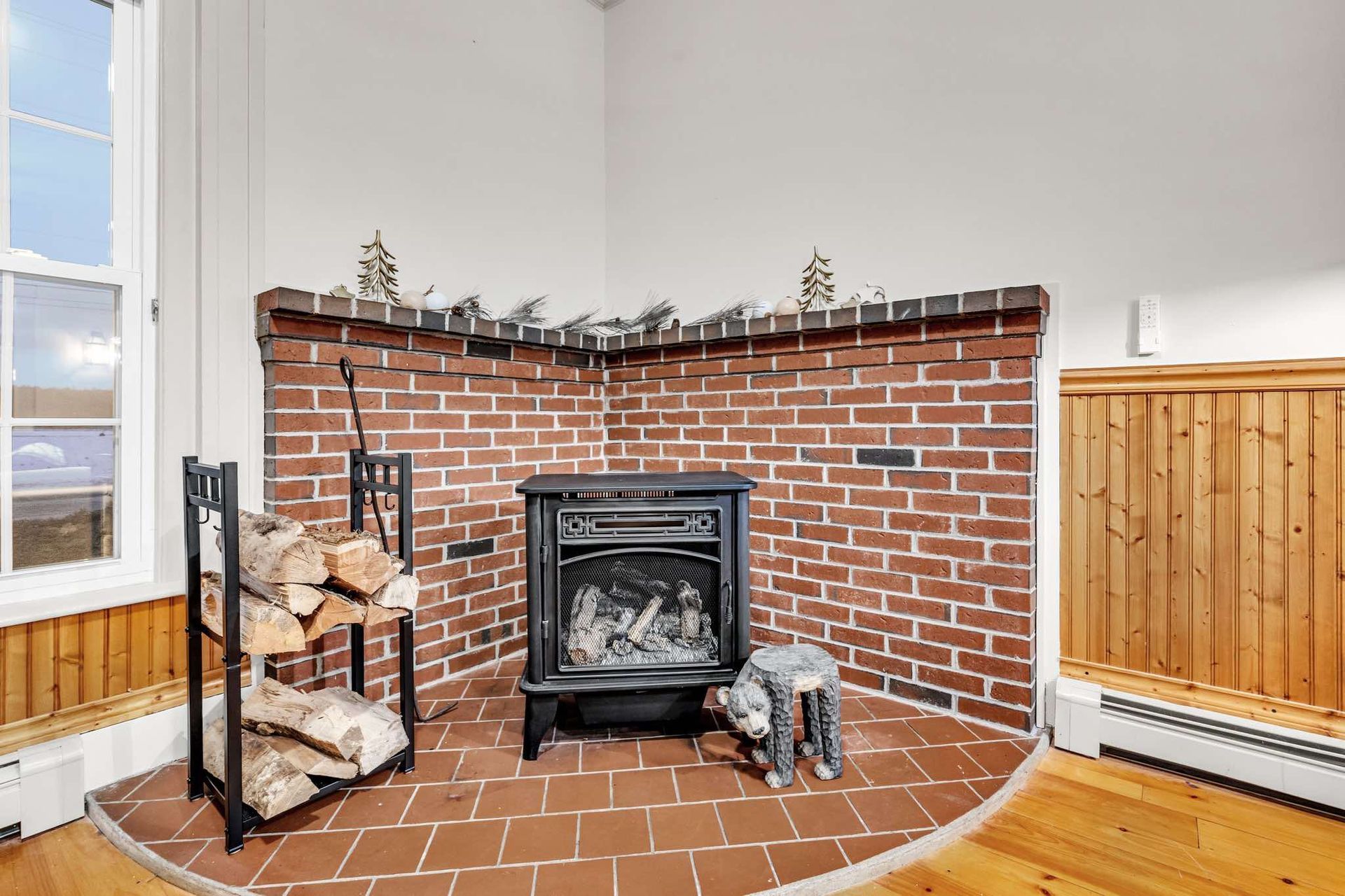 A brick fireplace with a black stove, firewood rack, and dog statue. Window on the left, wood paneling on the right.